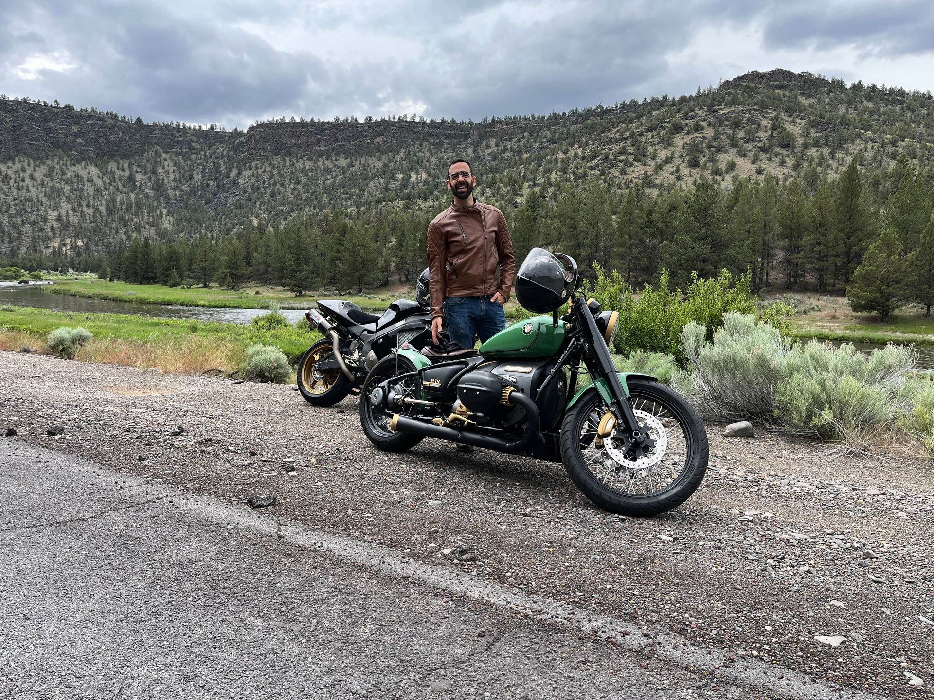 Person standing beside a black motorcycle on a roadside with mountains and trees in the background