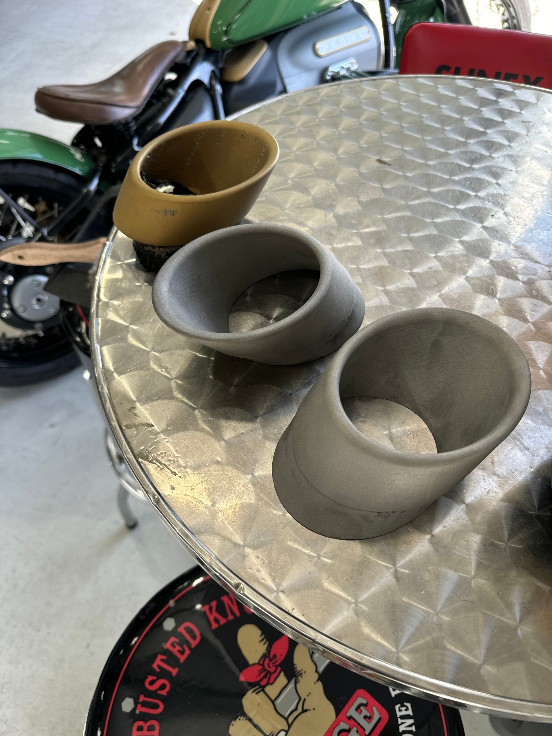 Three small cups on a silver diamond-plate table beside a motorcycle.