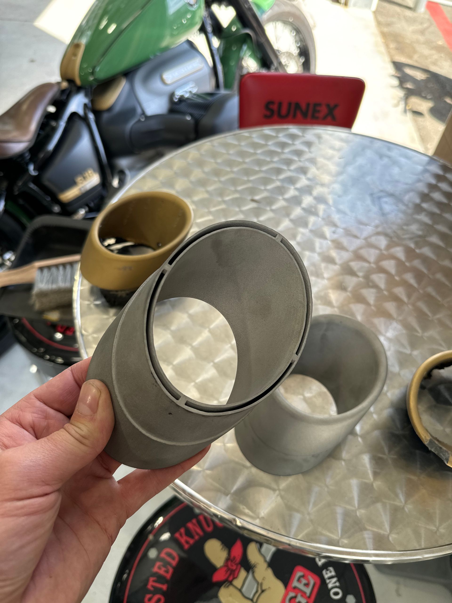 Hand holding a gray metal cylinder beside a clear round table with motorcycle parts and a green bike in the background
