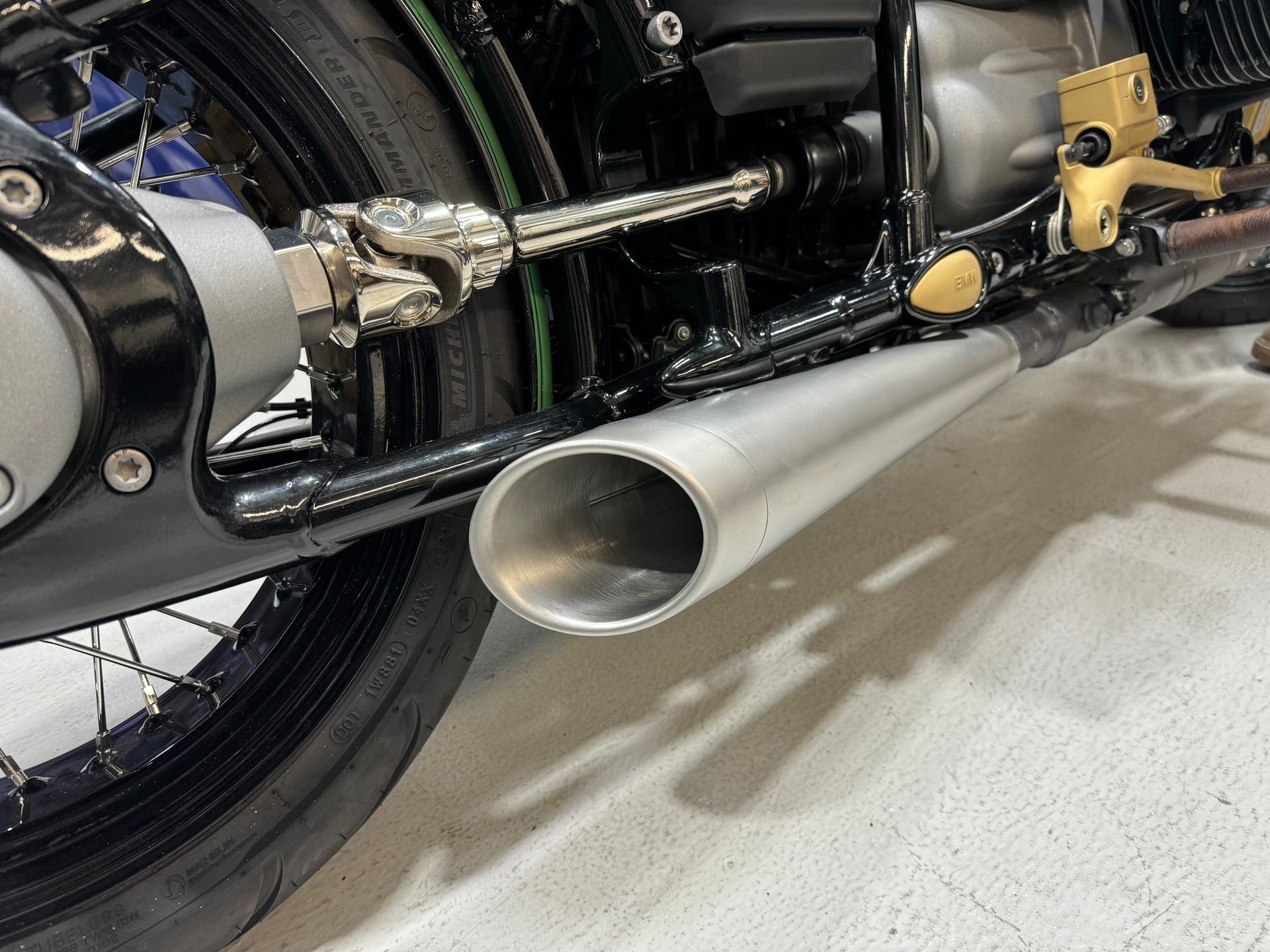 Close-up of a motorcycle’s chrome exhaust pipe, rear wheel, and foot peg on a light floor.