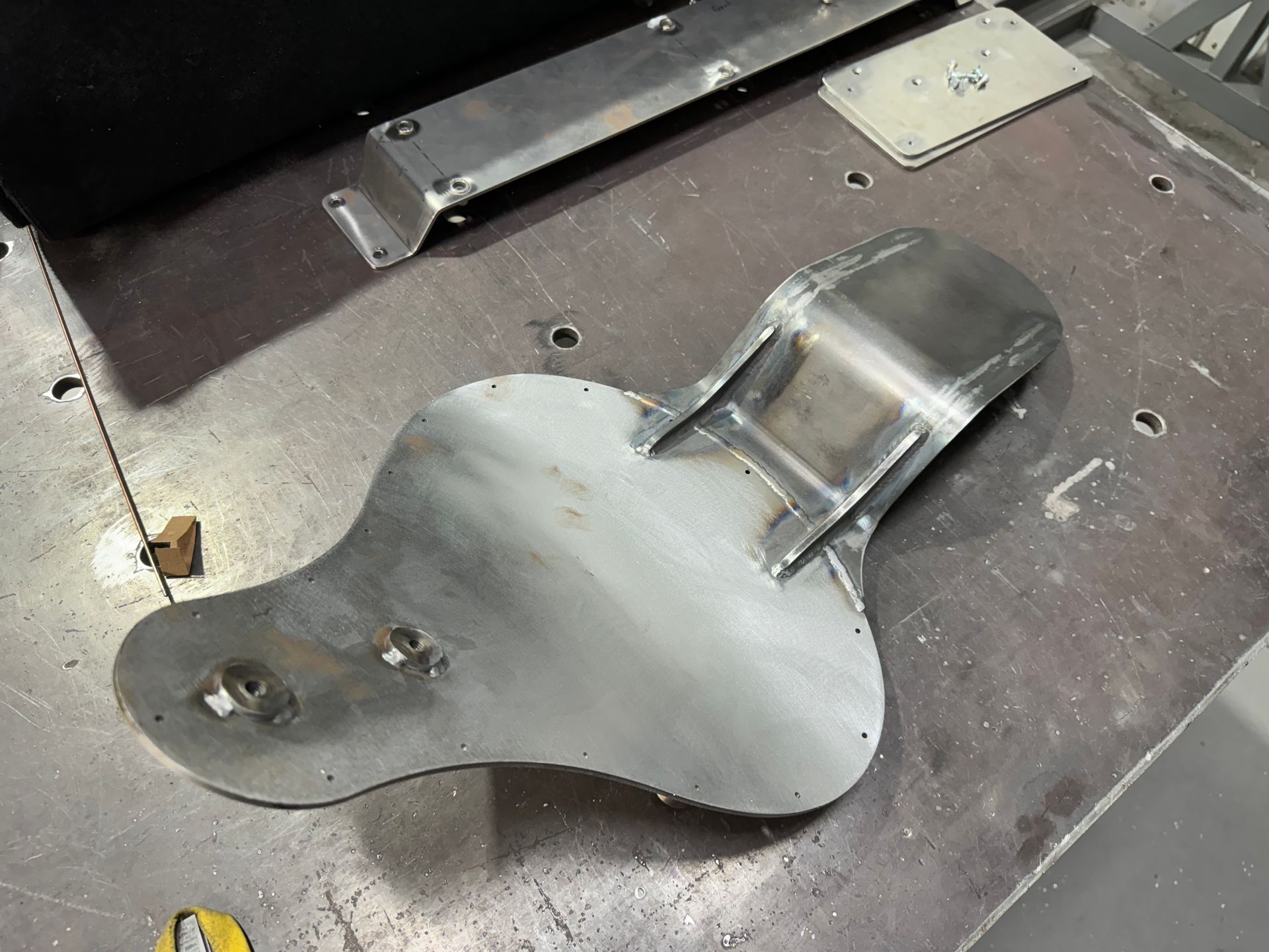 Metal motorcycle seat pan and backrest on a workshop floor, with bolts and a metal bracket nearby