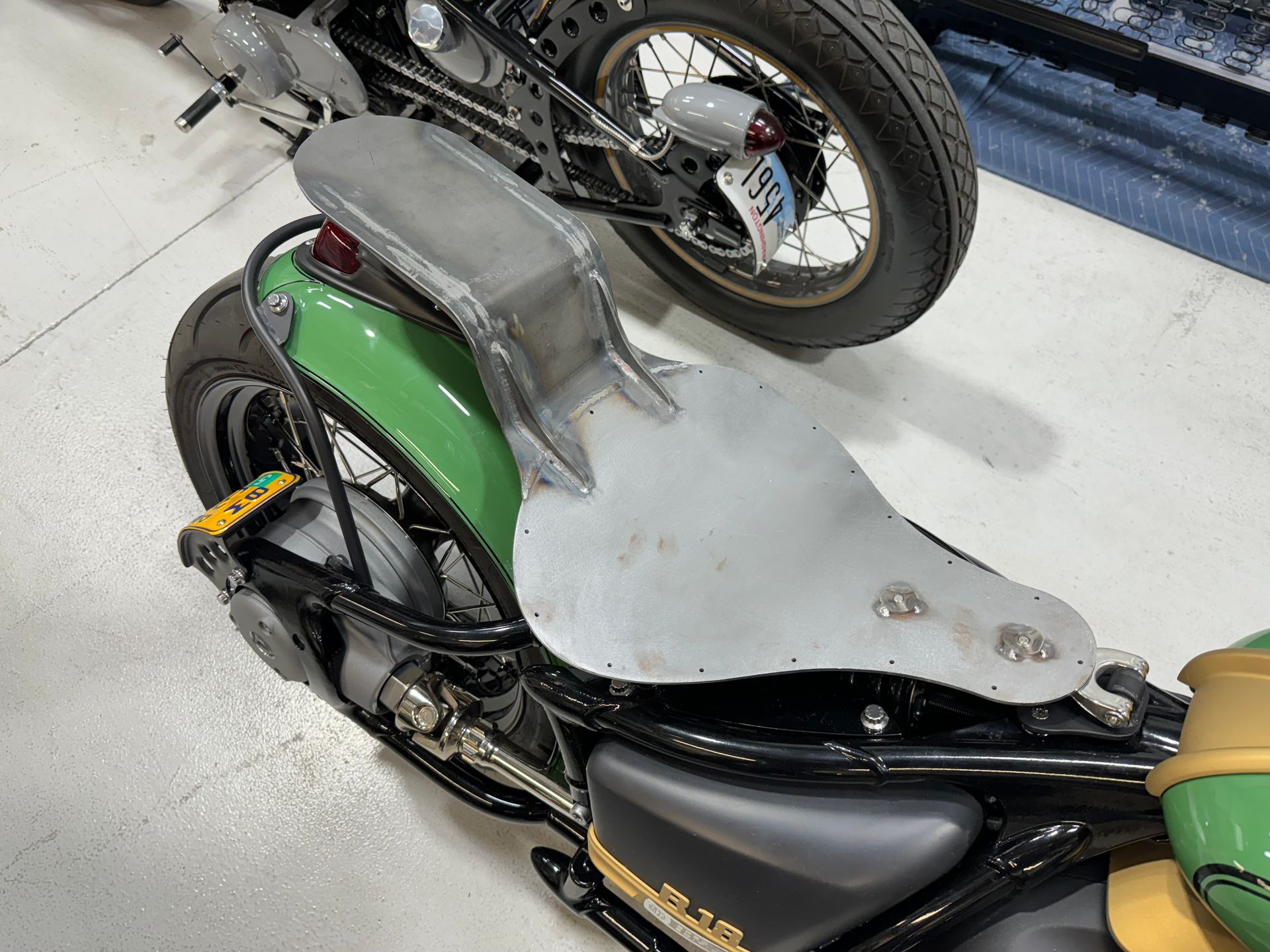 Two small motorcycles, one green and black, stacked on a workshop floor with a large silver panel between them