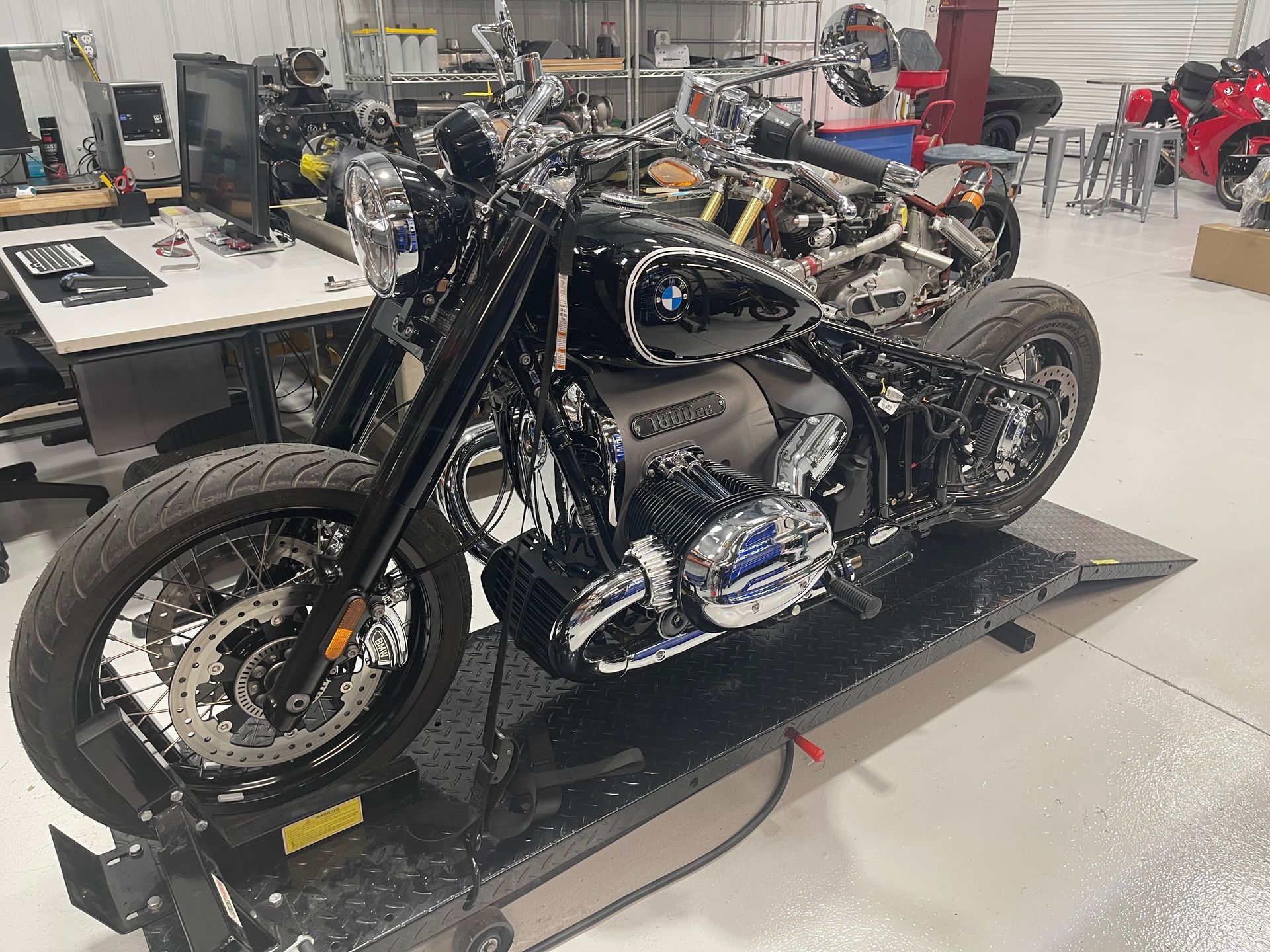 Black cruiser motorcycle on a platform in a workshop, with chrome details and other bikes in the background