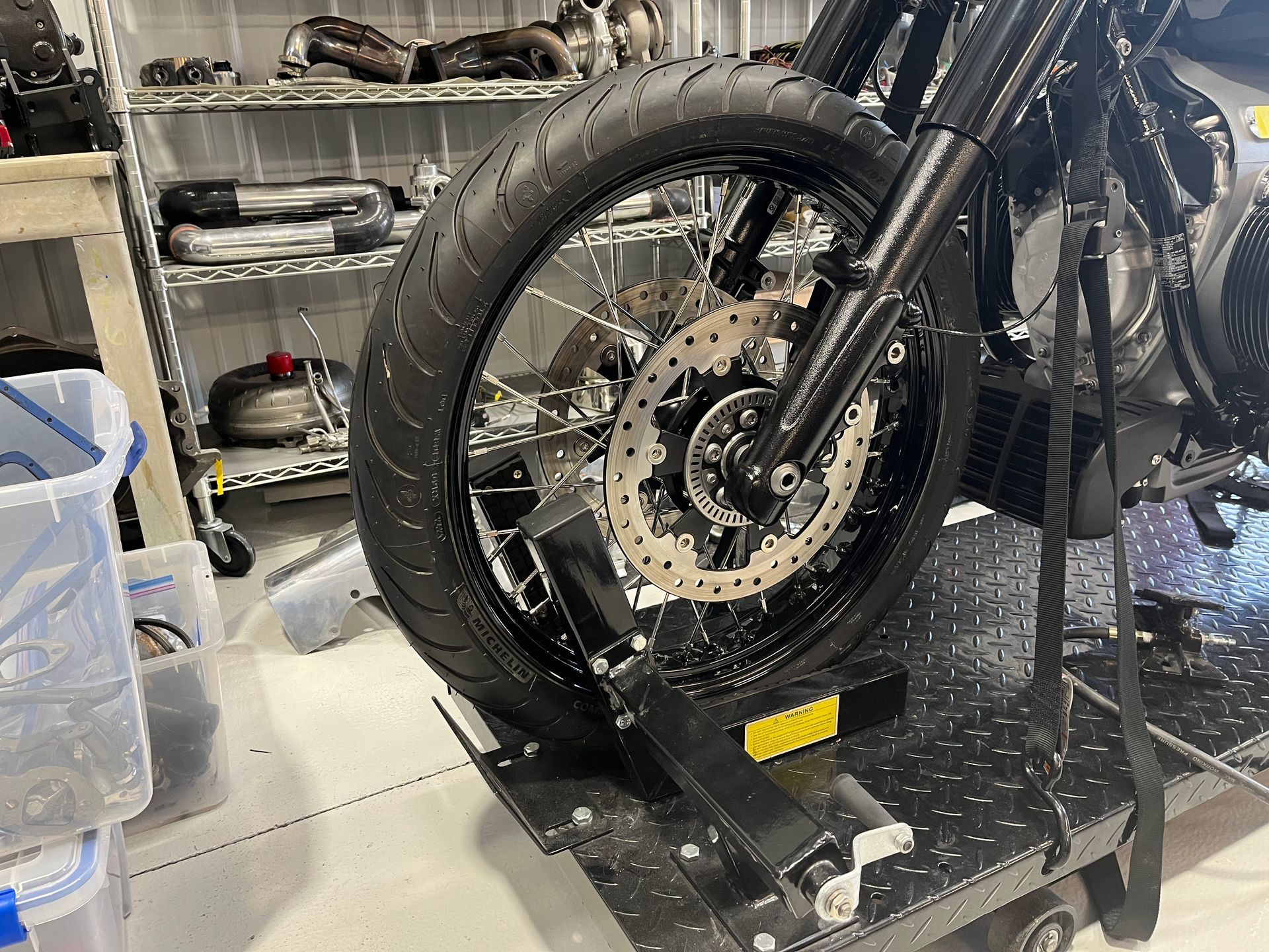 Motorcycle front wheel on a maintenance stand in a garage, with tools and parts shelves behind it