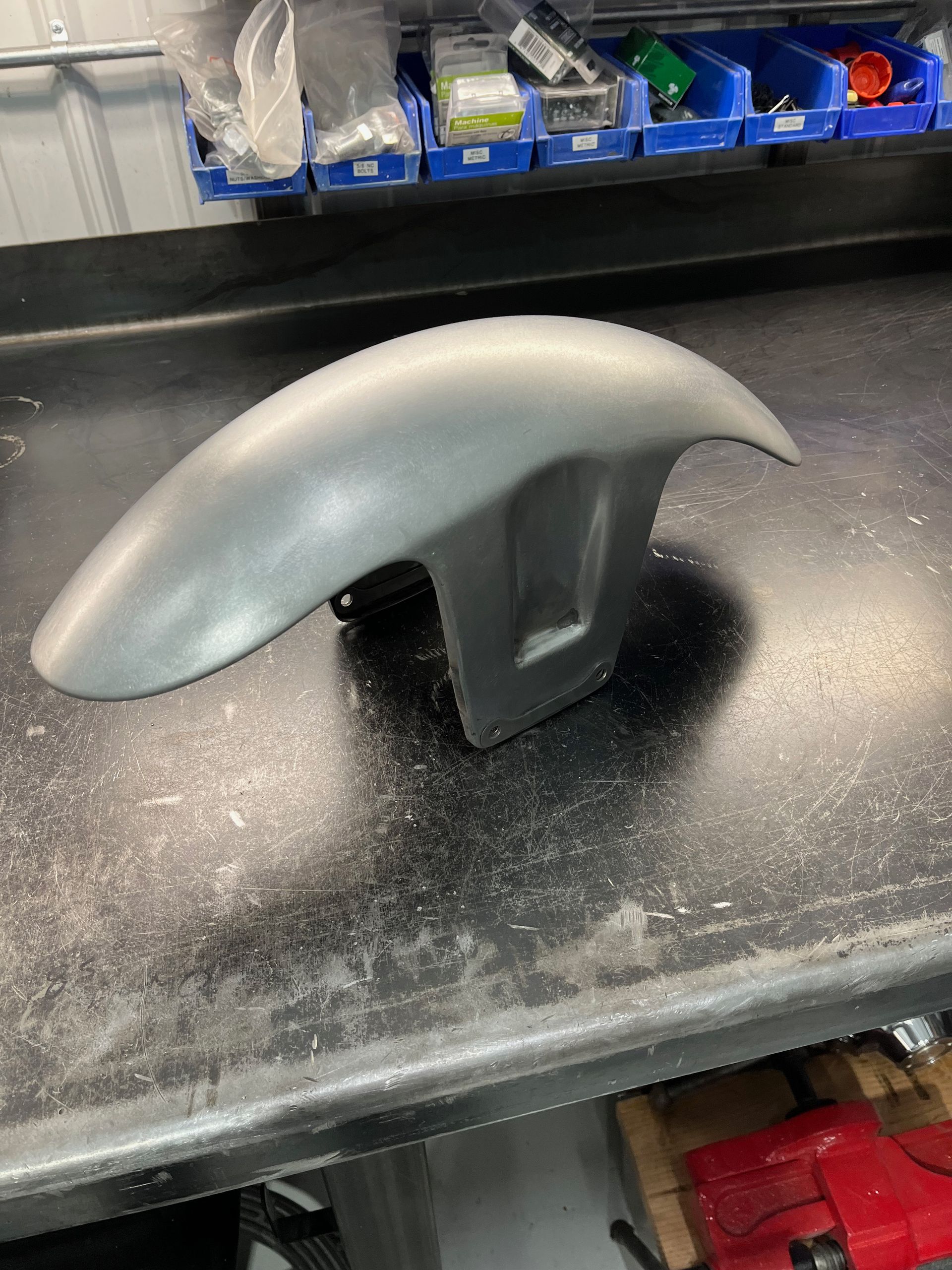 Silver curved object on a black workbench in a workshop, with blue bins in the background