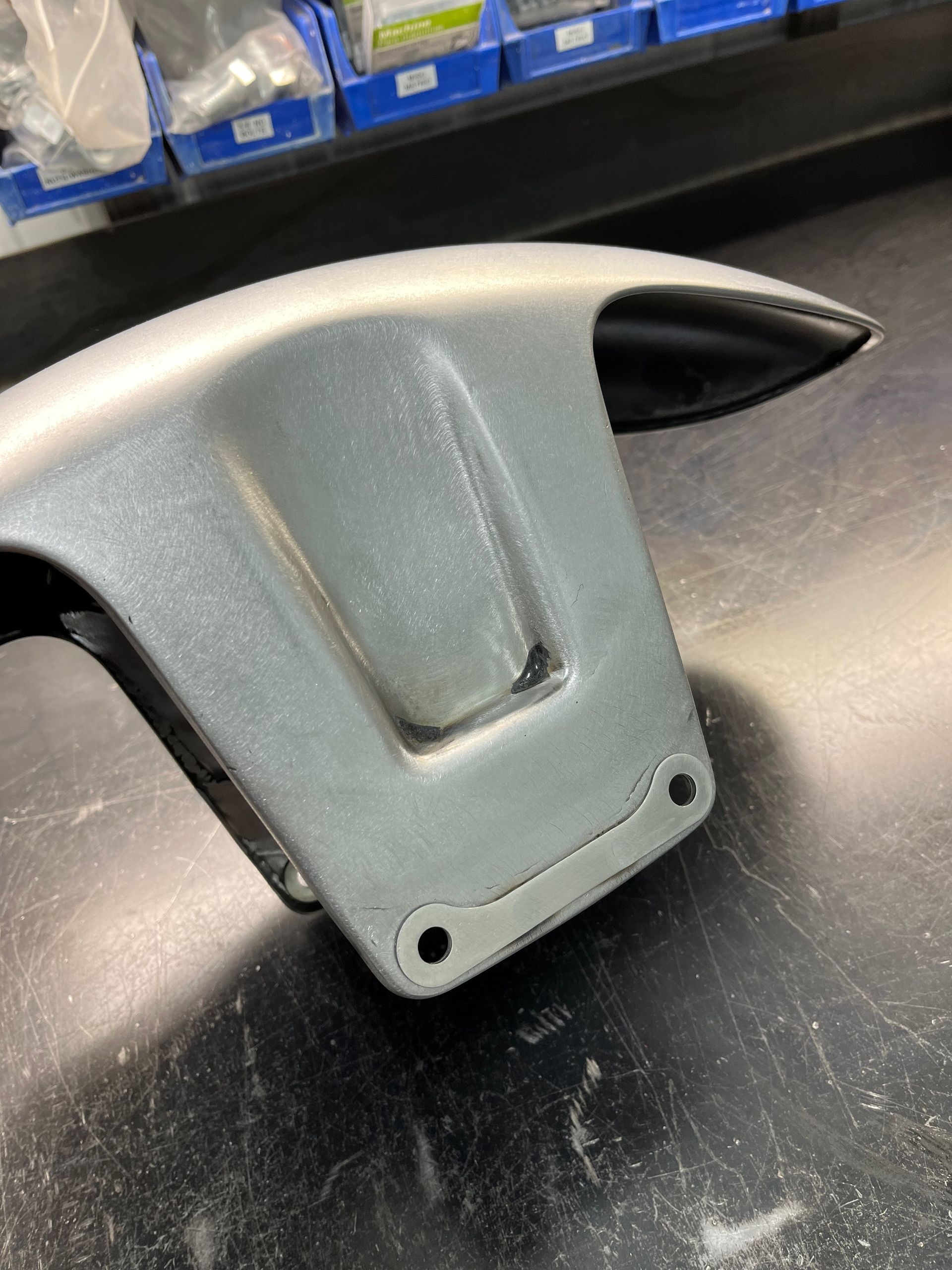Gray plastic bracket on a black surface, with two holes at the bottom and shelves in the background