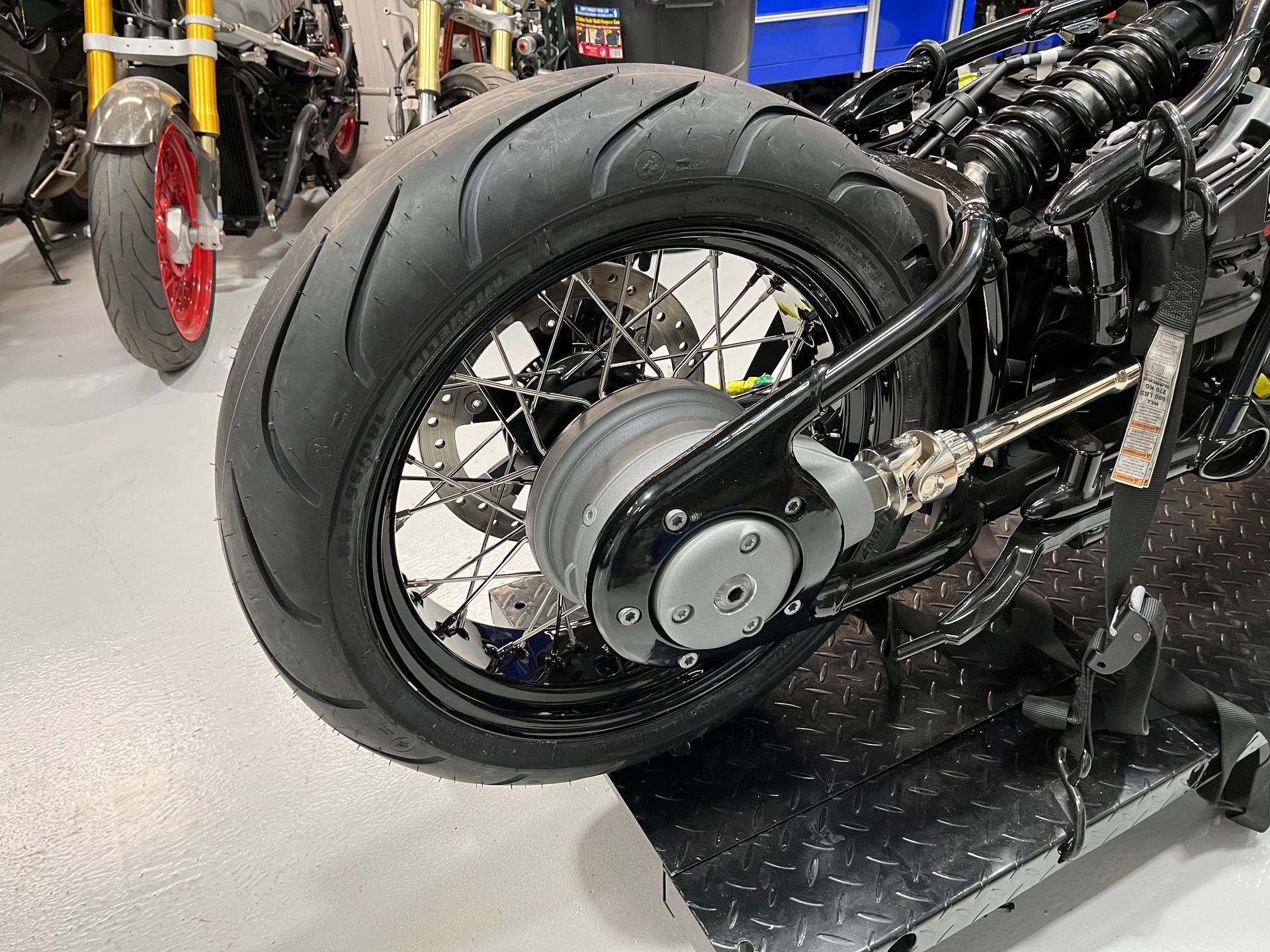 Black motorcycle rear wheel and chain assembly in a workshop, with a thick tire and spoked rim.