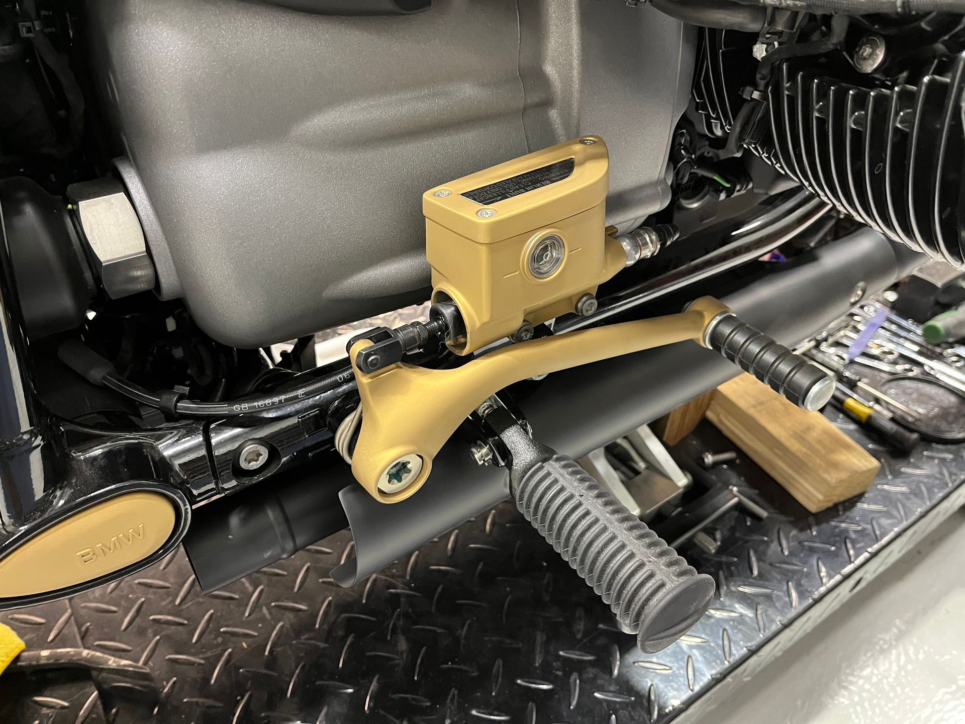 Gold motorcycle foot control on black diamond-plate floor, with engine and foot peg visible