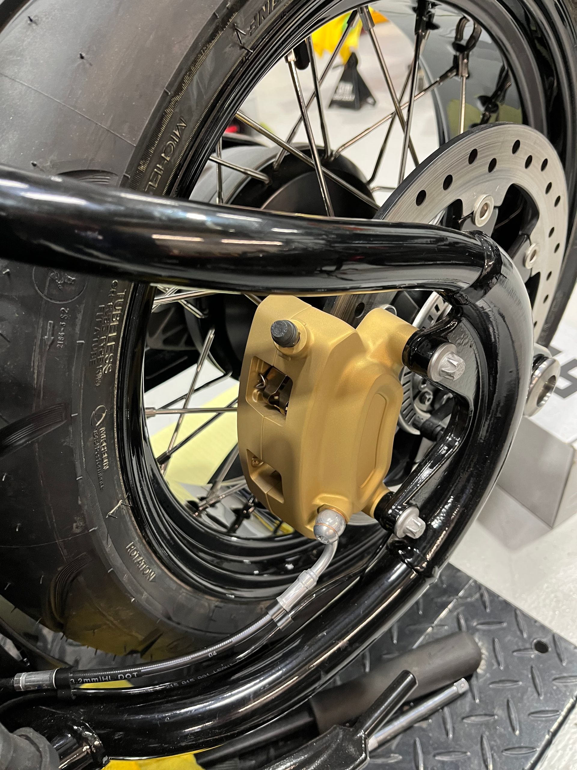 Close-up of a motorcycle wheel with a black tire, disc brake, and yellow brake caliper in a workshop.