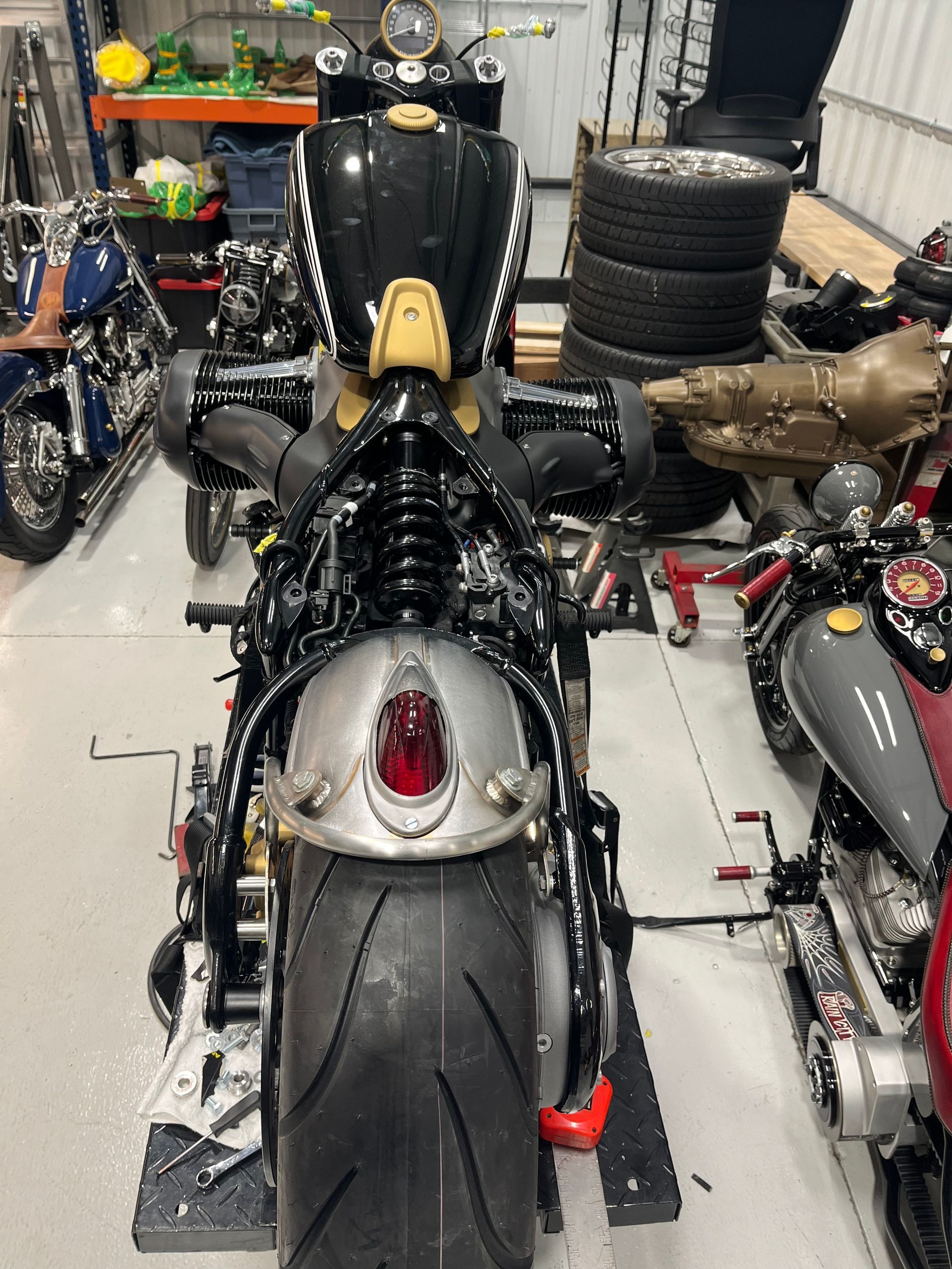 Two custom motorcycles in a workshop, with a black bike centered in the foreground and parts stacked behind.