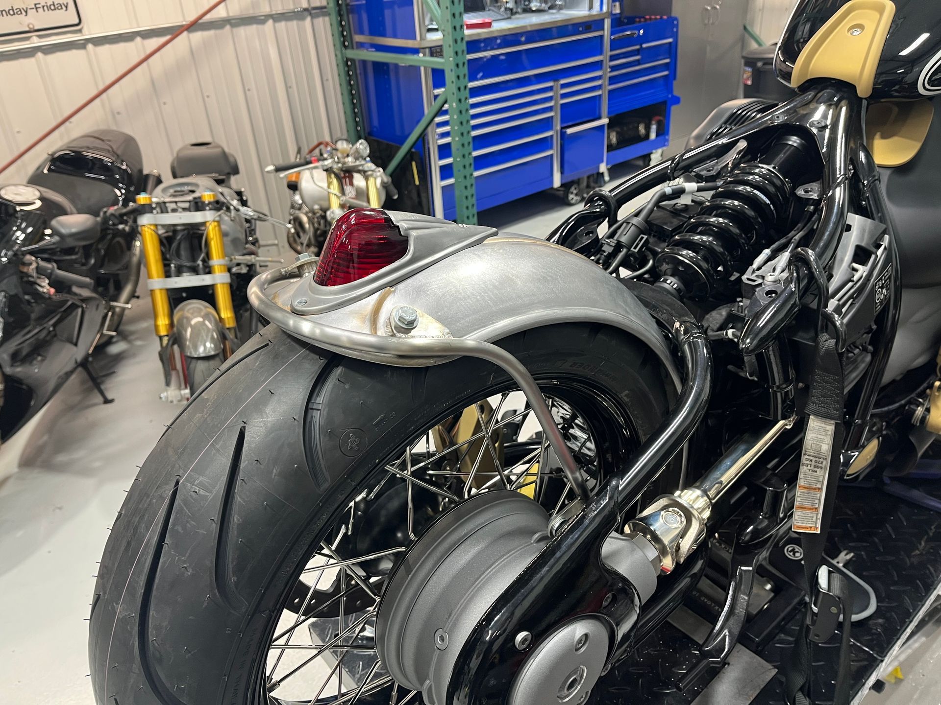 Custom motorcycle in a workshop, showing a wide rear tire, exposed frame, and blue tool chest in the background.