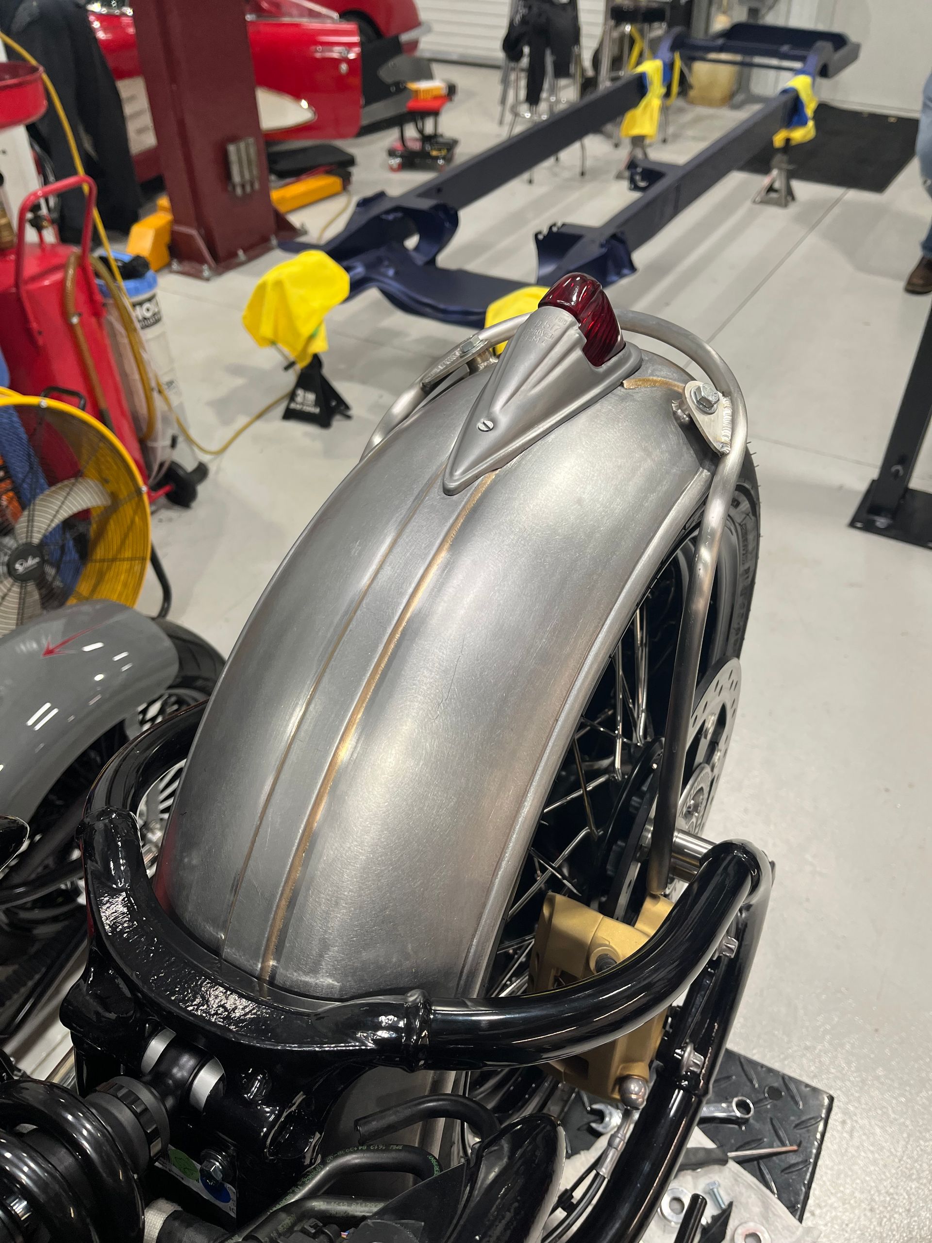 Silver vintage car body on a vehicle lift in a garage, with black chassis and yellow wheel chocks.