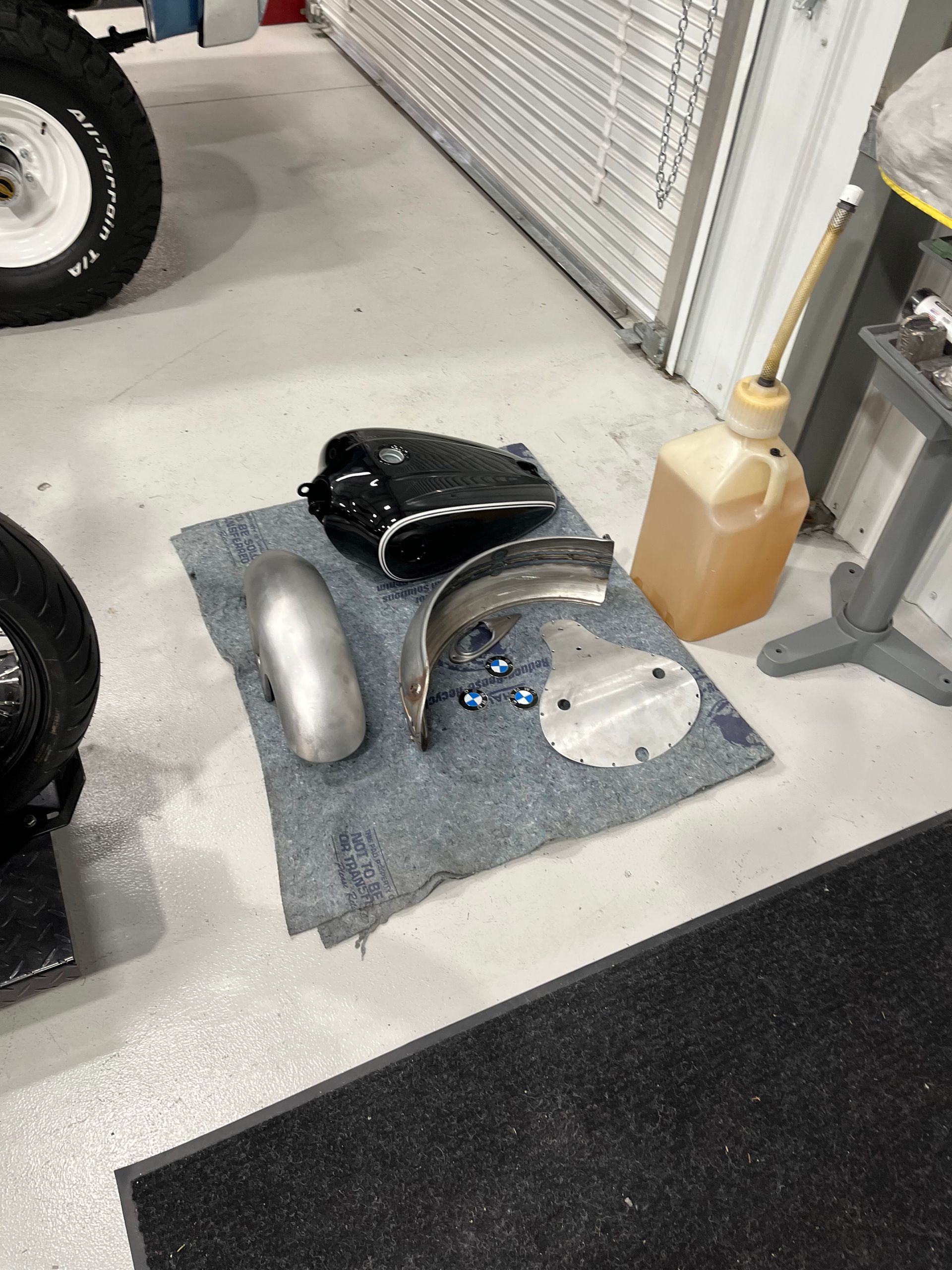 Car parts laid out on a floor mat in a garage, including a black fender, silver panel, and blue hardware.