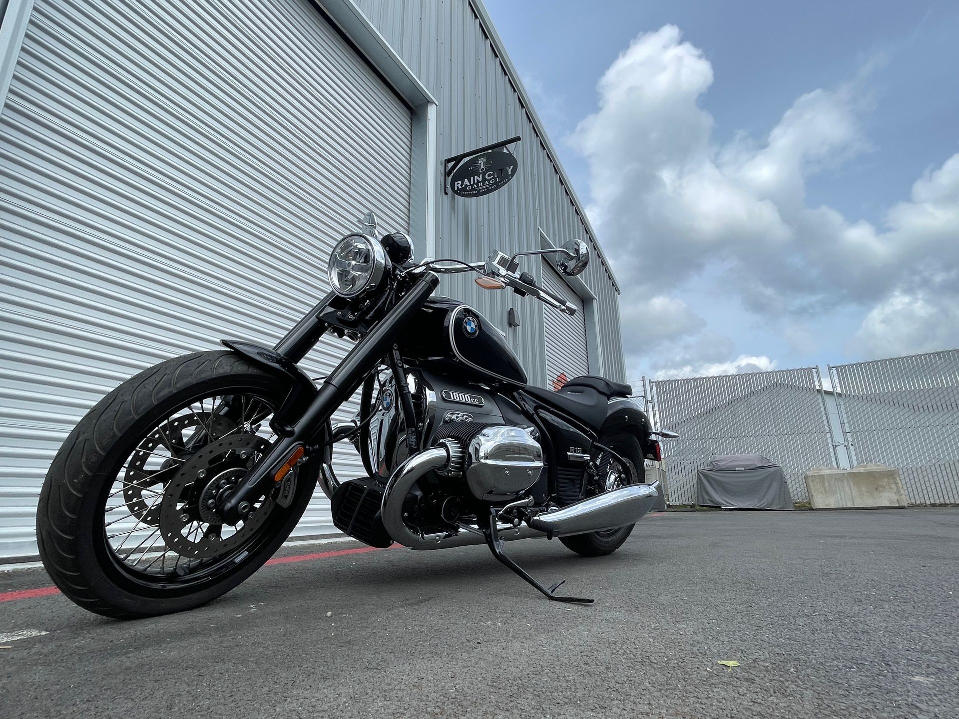 Black cruiser motorcycle parked beside corrugated metal buildings under a cloudy sky.