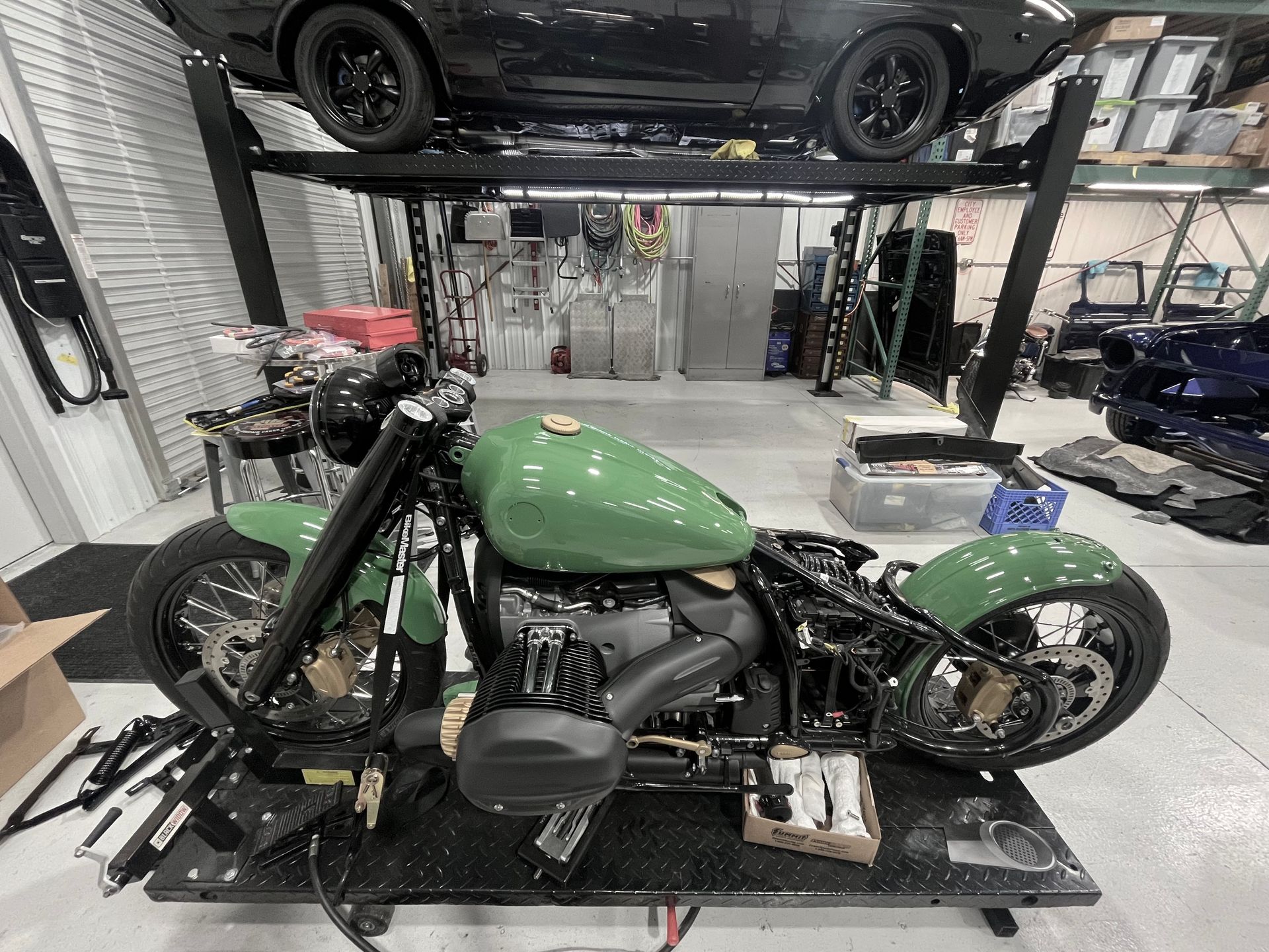 Green custom motorcycle on a lift in a workshop, with another car elevated overhead.