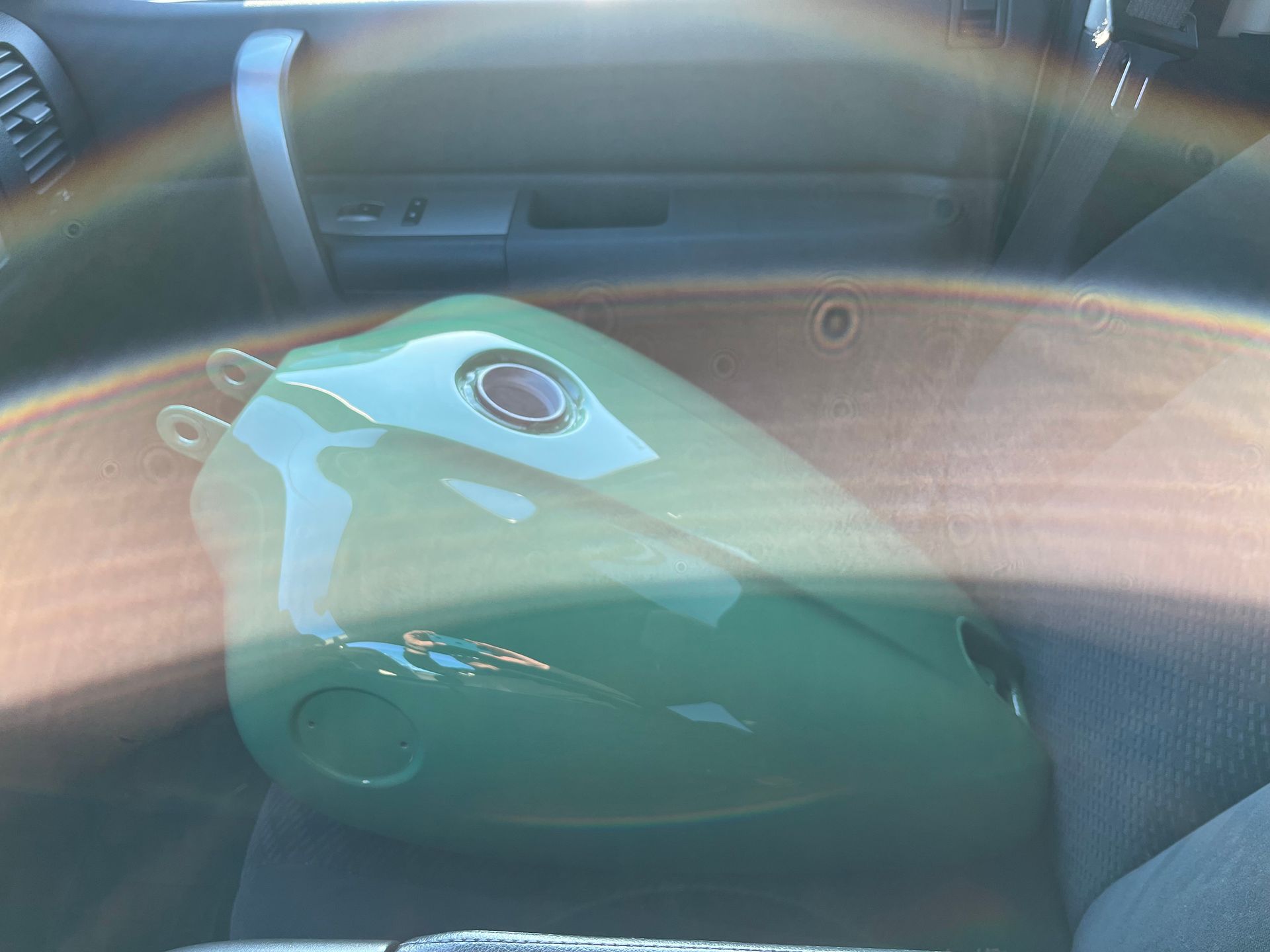 Green motorcycle fuel tank on a seat inside a car, viewed through a reflective window.