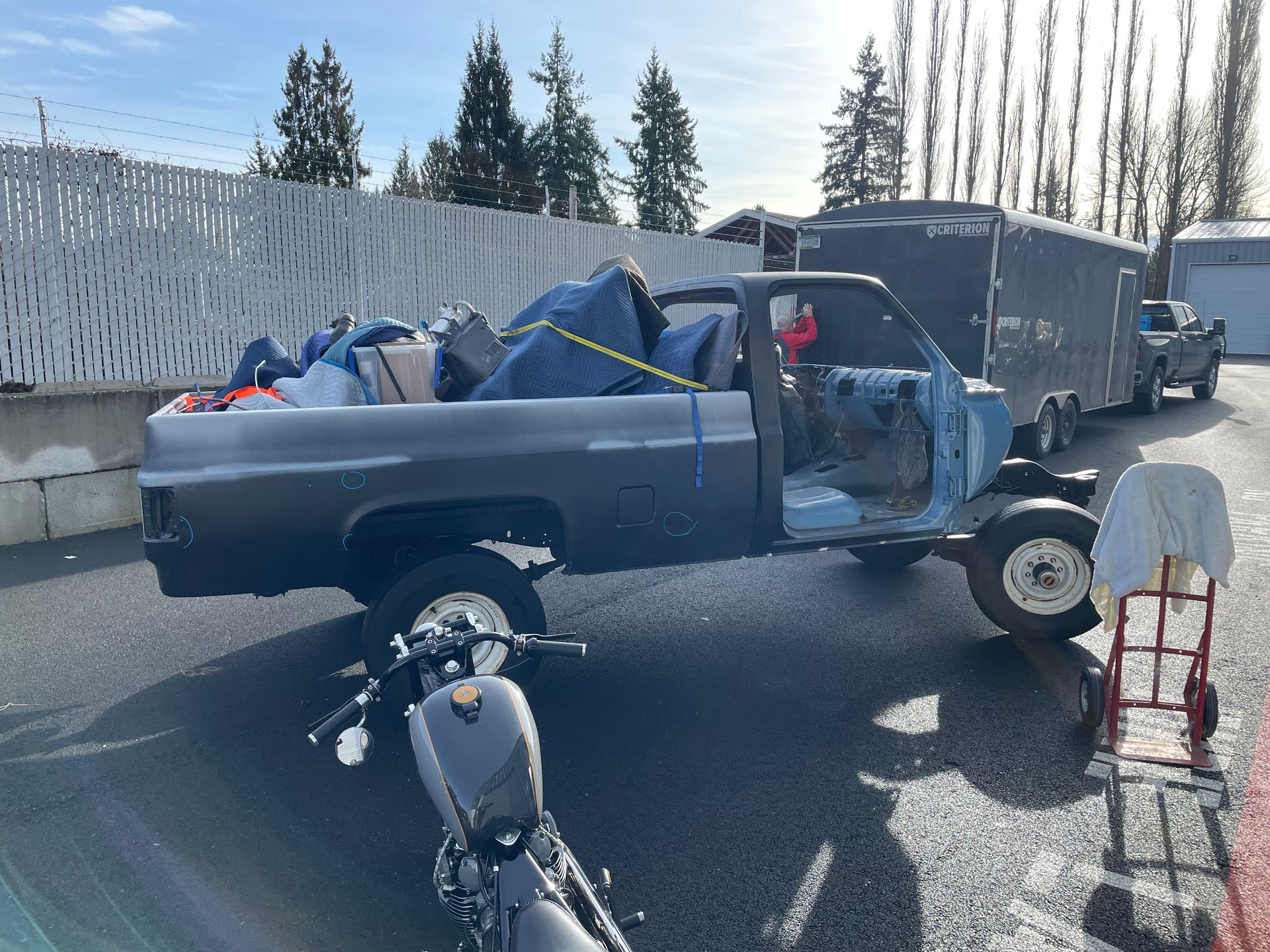 Blue pickup truck loaded with bags and debris at a roadside, with a tow trailer and scooter nearby.
