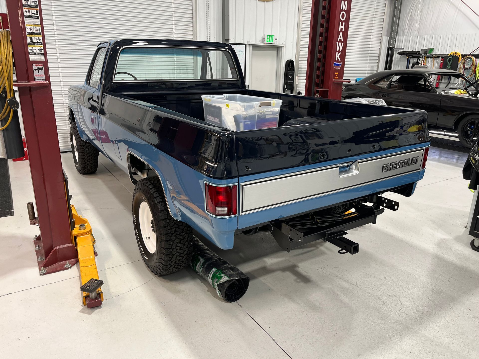 Blue classic pickup truck on a lift in an auto shop, rear view with open bed and exhaust visible