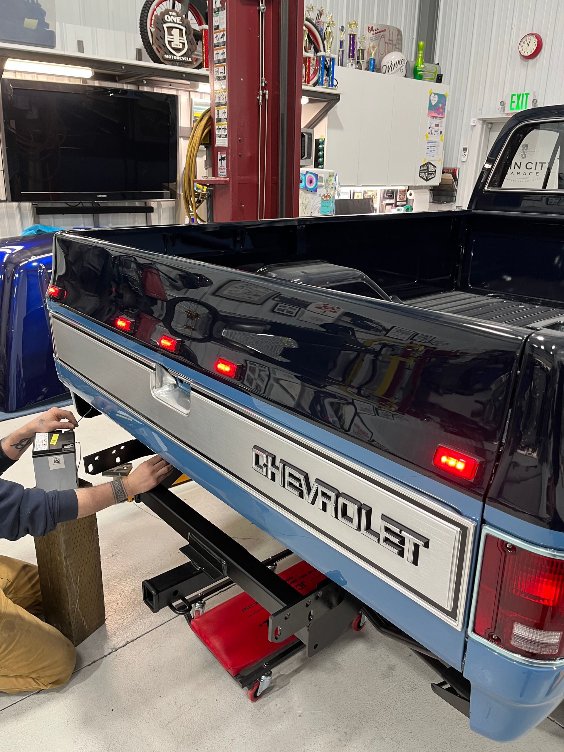 Blue Chevrolet Avalanche on a red lift in a garage, rear view with open bed and lit tail lights.