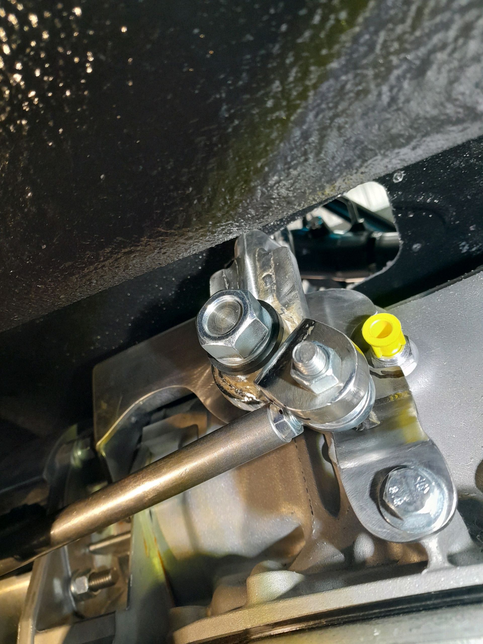 Close-up of a metal mechanical linkage with bolts and a yellow cap under a dark surface.