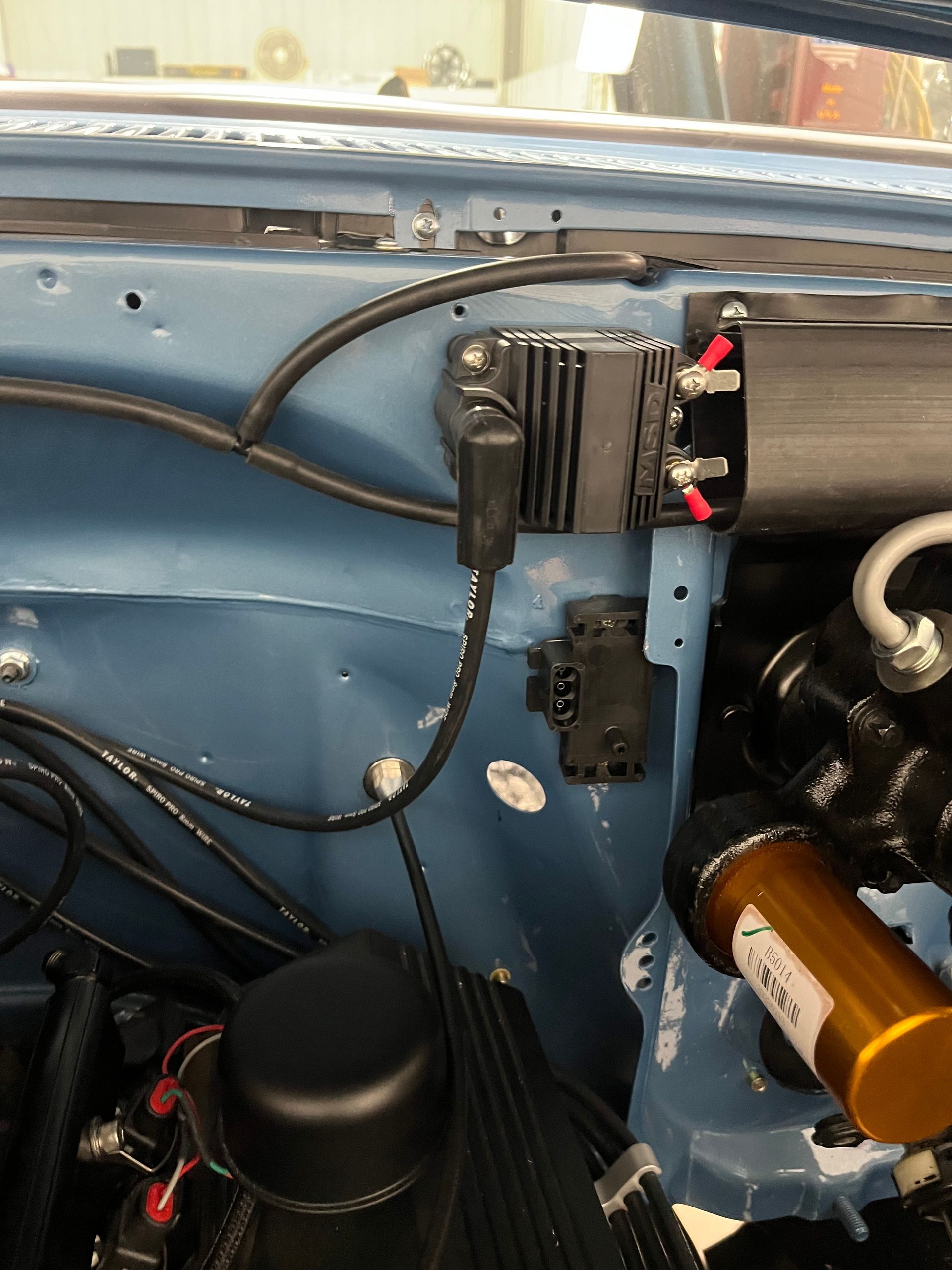 Blue car interior with wiring, a black electronic module, and a copper-colored cylinder mounted near the engine bay.