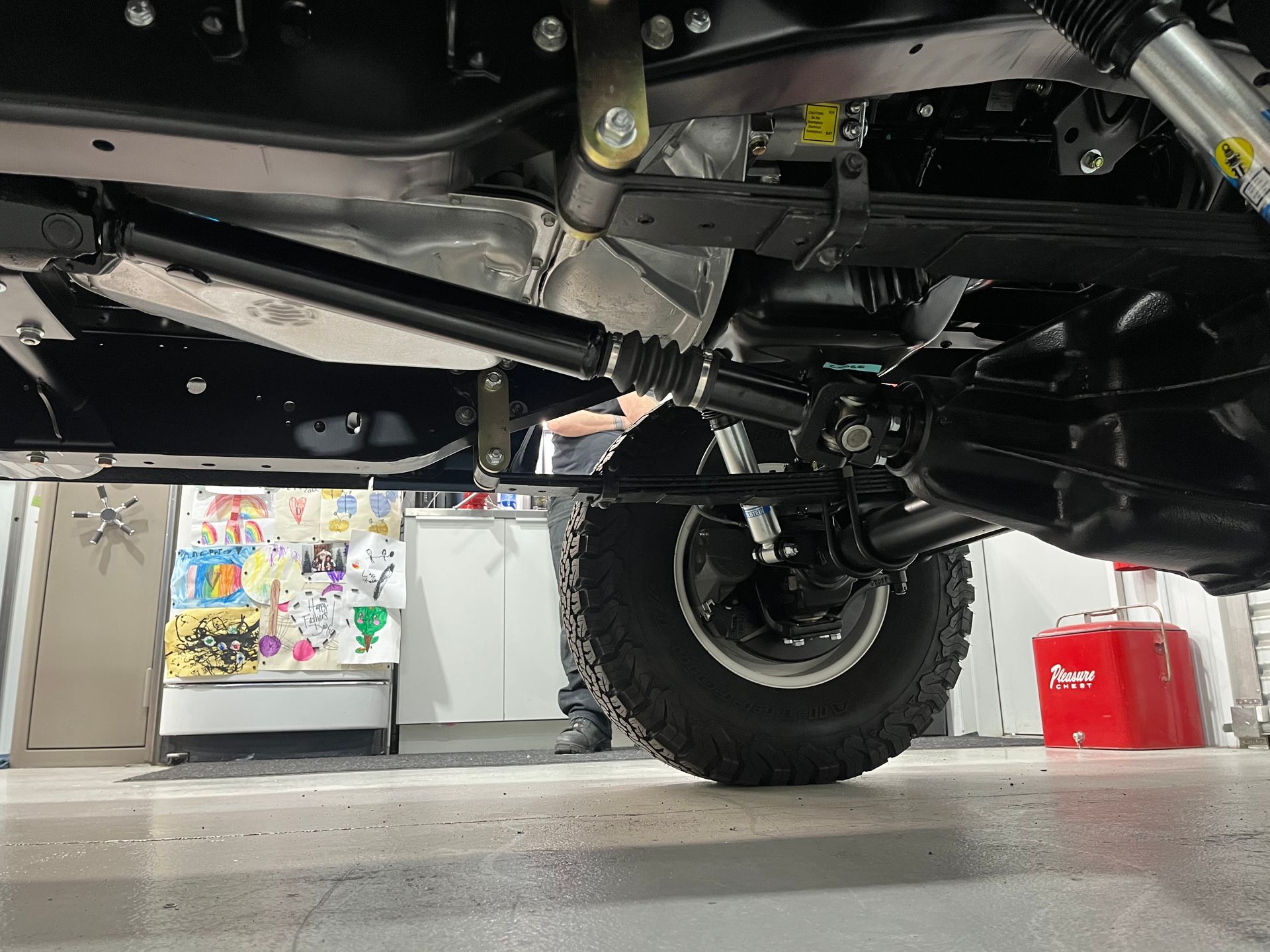 Undercarriage of a black off-road truck, showing suspension and rear tire in a garage.