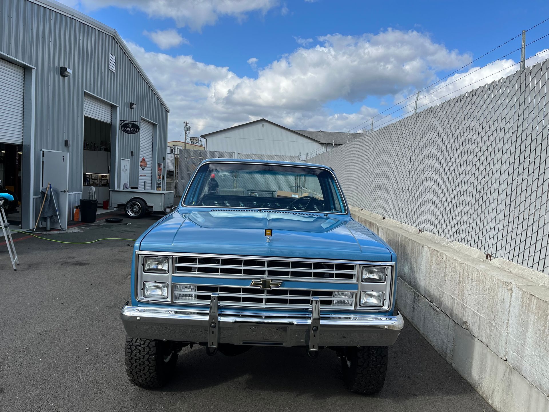 Blue vintage pickup truck parked beside a warehouse and chain-link fence.