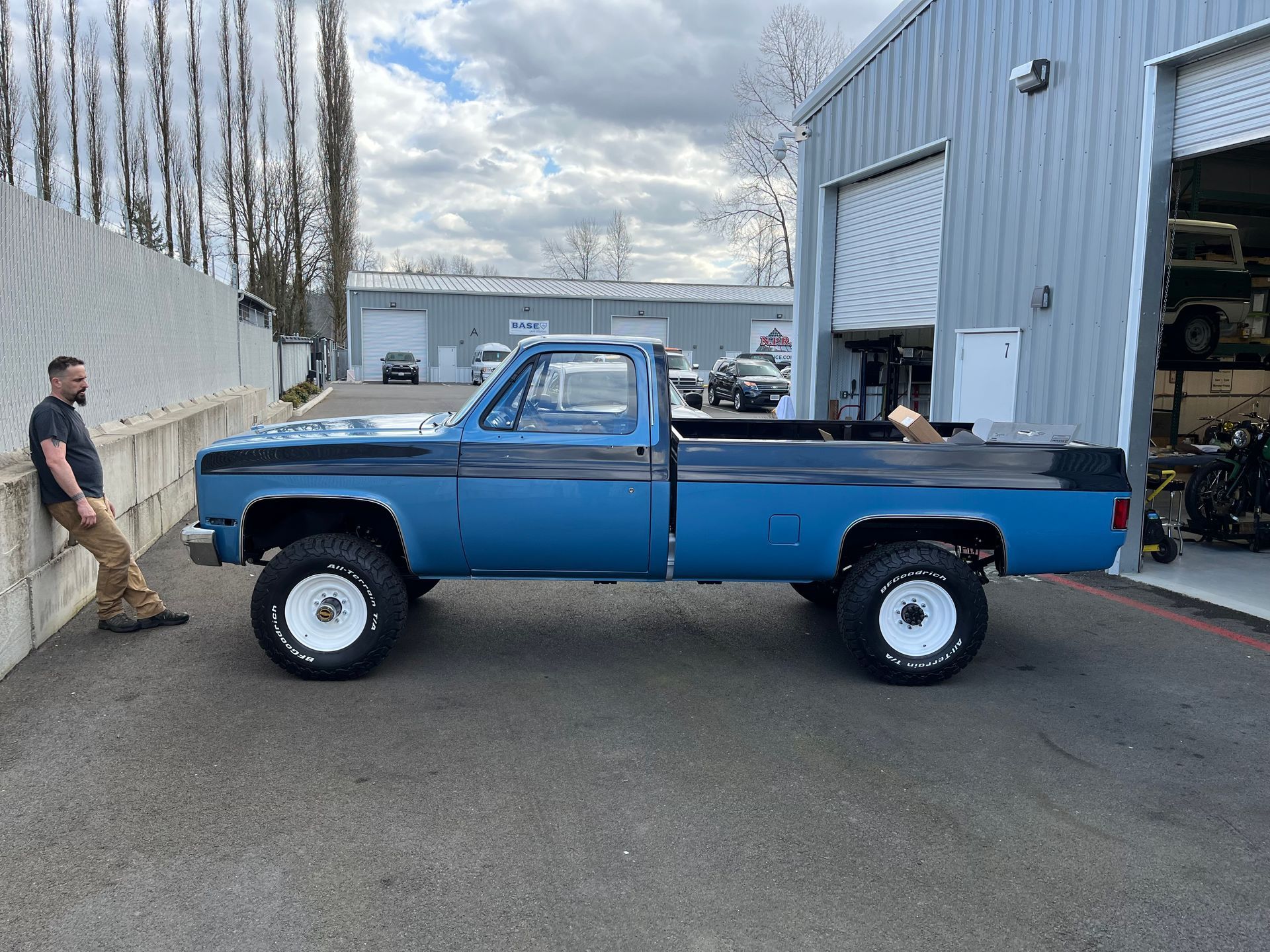 Blue lifted pickup truck parked outside a garage, with a man standing nearby.