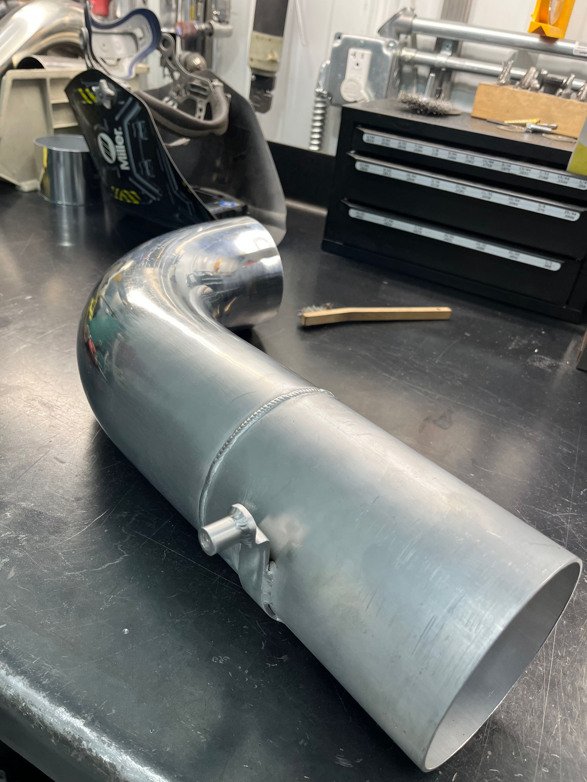 Large silver metal pipe section on a workbench in a workshop, with tools and a parts drawer in the background