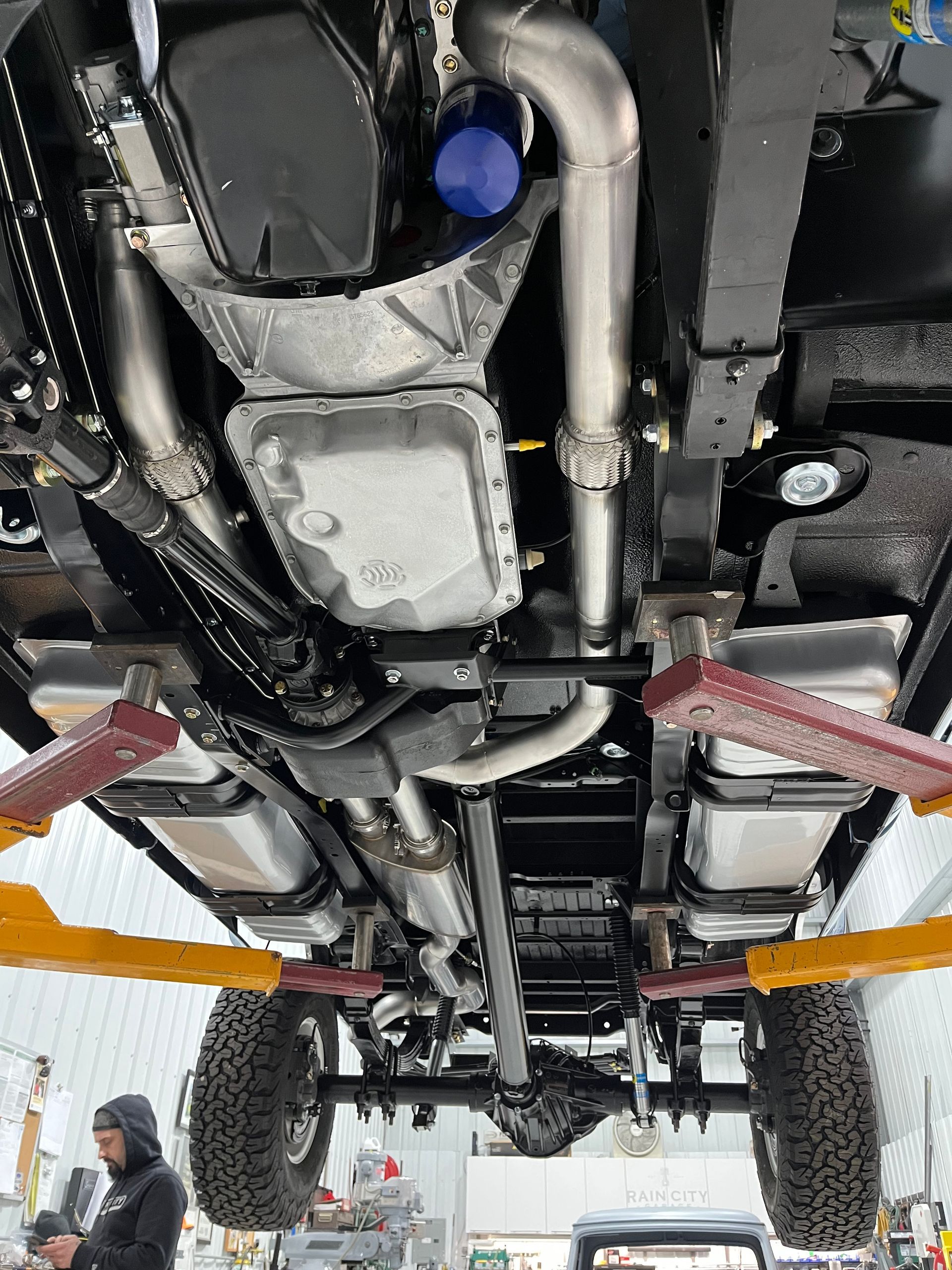 Underside of a lifted vehicle, showing exhaust, suspension, and large off-road tires in a workshop.