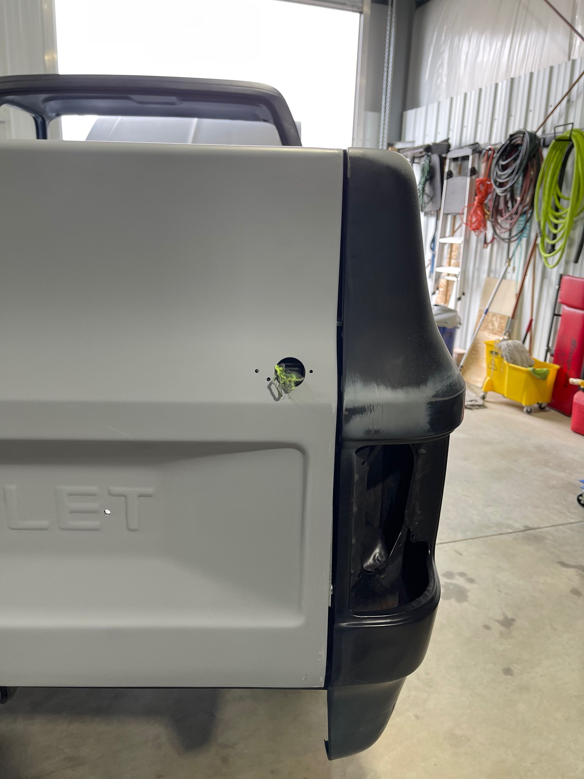 Rear side of a light gray Chevrolet truck in a garage, with a dark damaged rear corner.