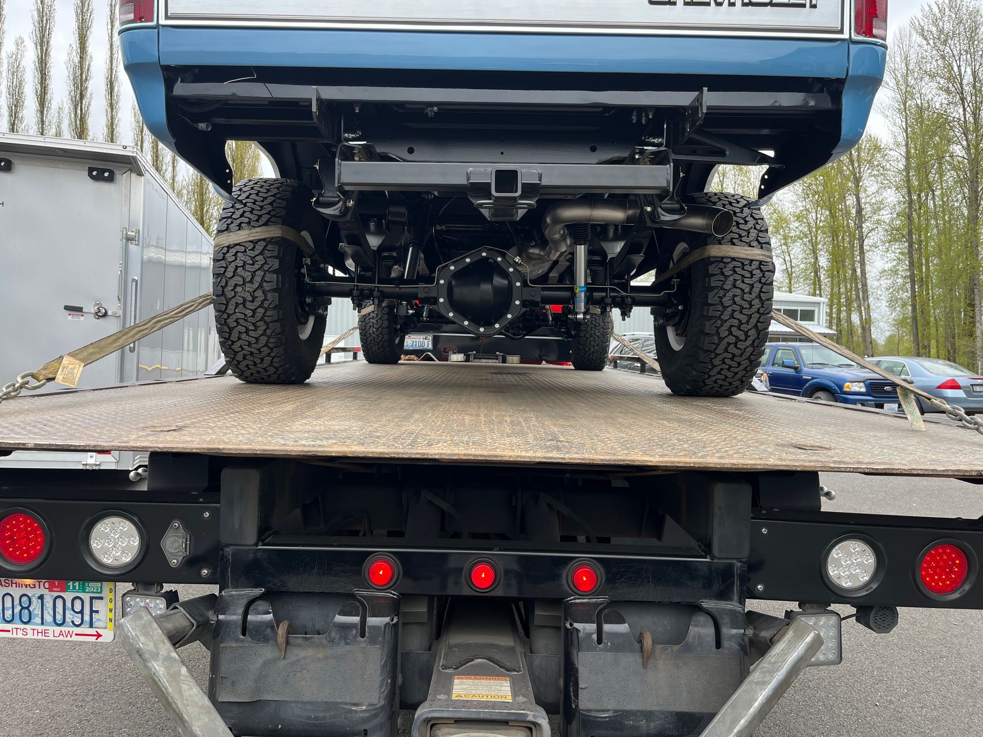 Blue pickup truck loaded on a flatbed tow truck, rear view on a road with trees in the background