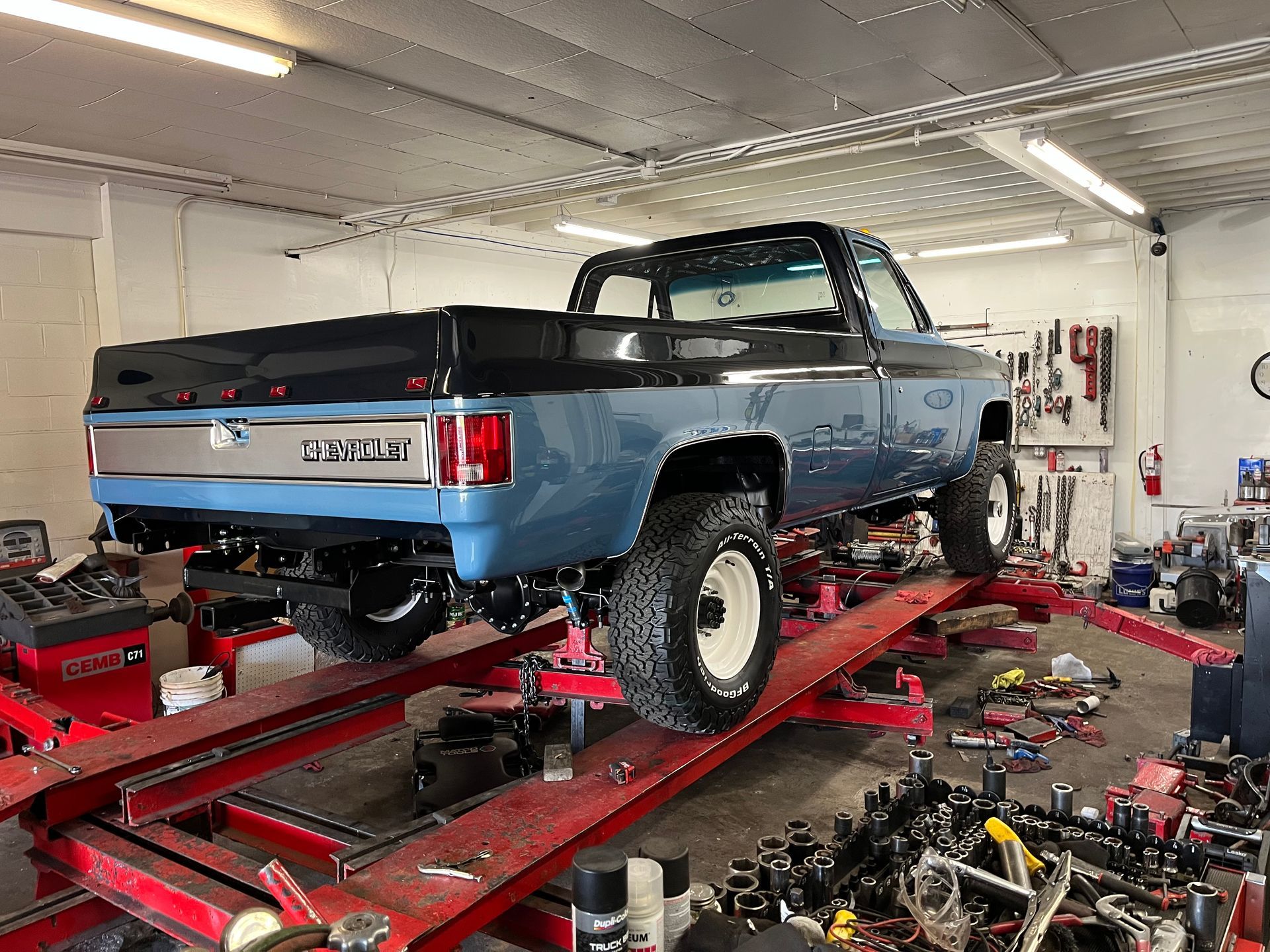 Blue classic pickup truck on a red lift in a cluttered auto repair shop with car parts scattered around
