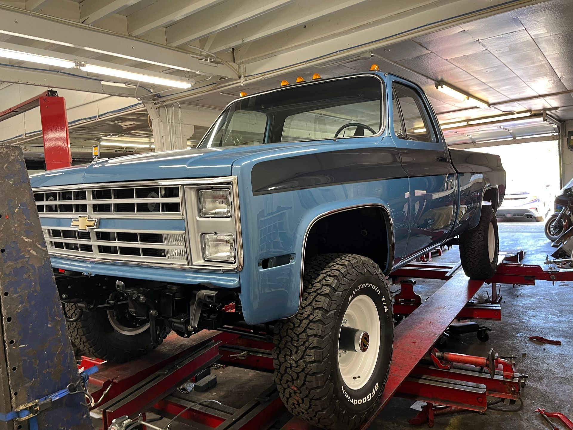 Lifted blue-and-black vintage truck in a repair shop, raised on a red lift with oversized tires.