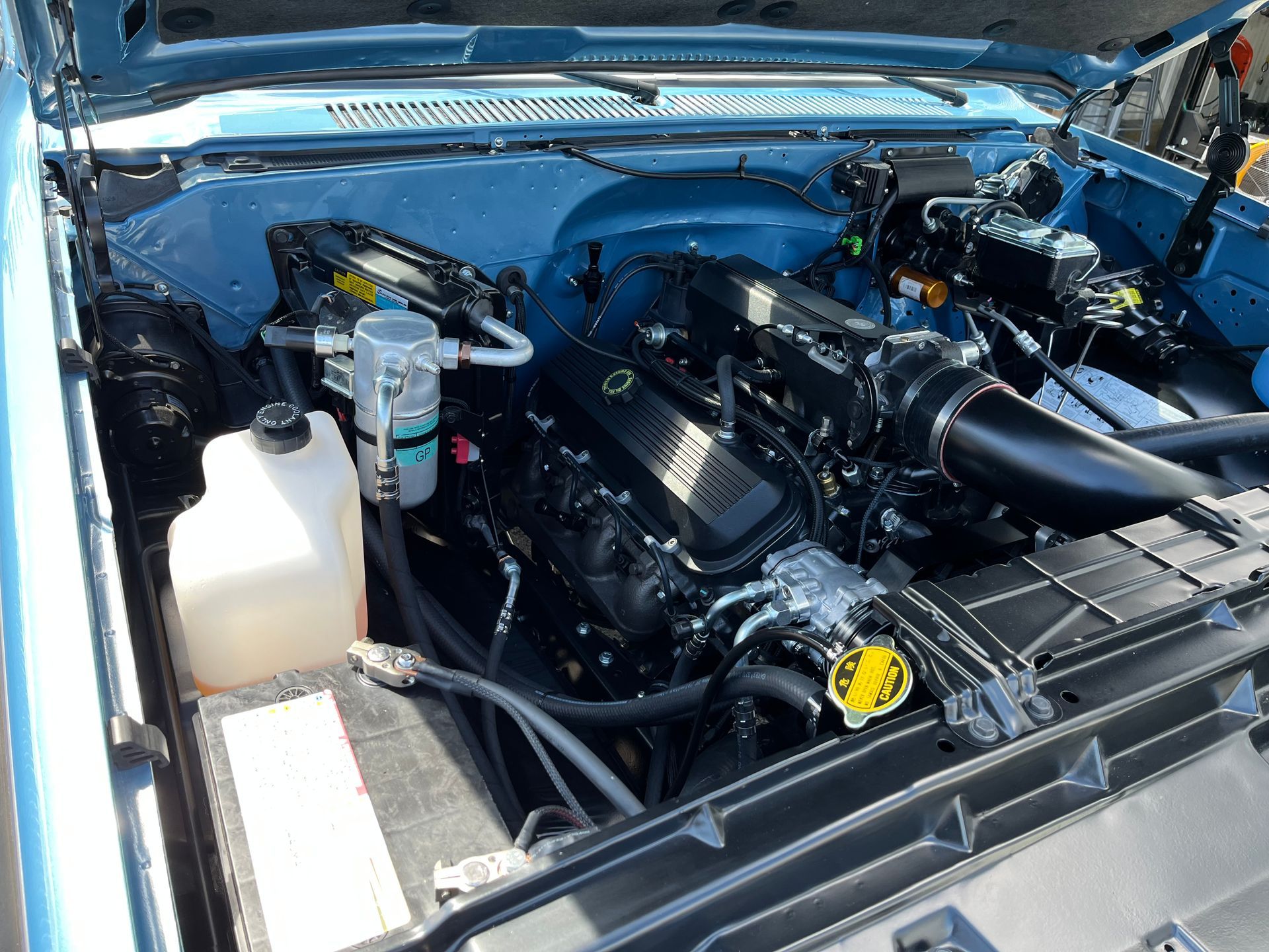 Blue car engine bay with black engine components and white coolant reservoir open under the hood