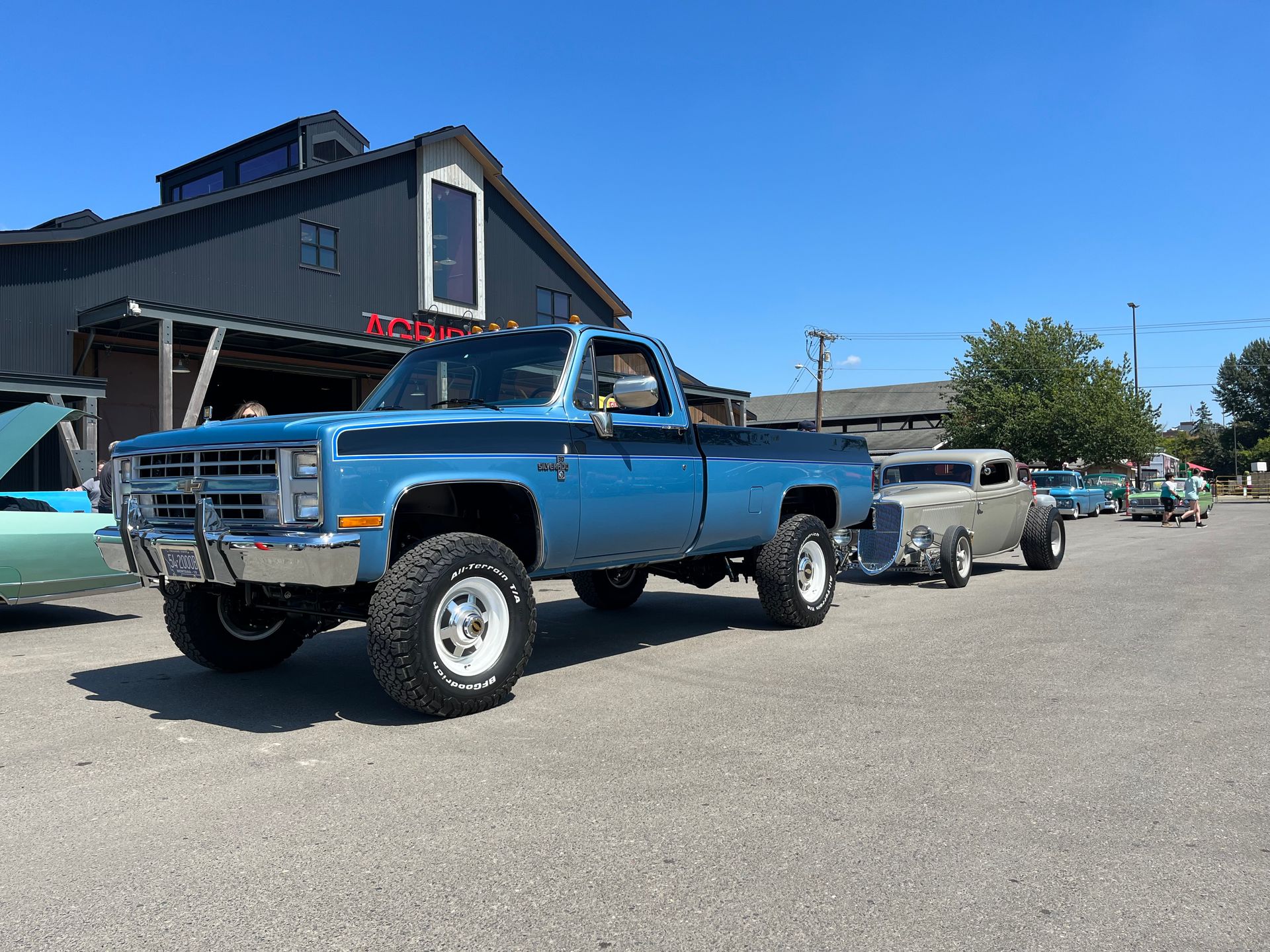 Blue lifted vintage pickup truck parked on a sunny street beside classic cars and a building