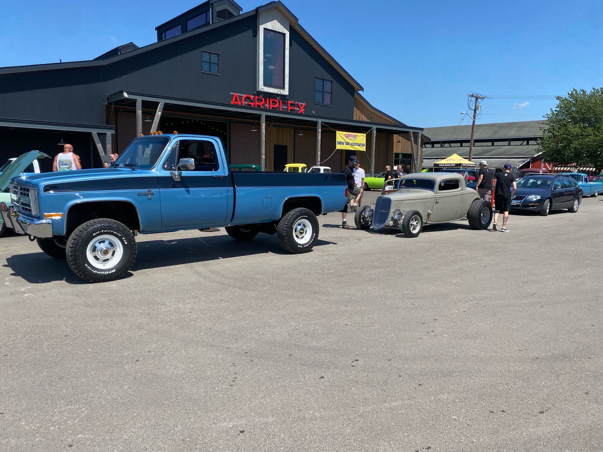 Blue pickup trucks and a gray hot rod parked outside a roadside building on a sunny day