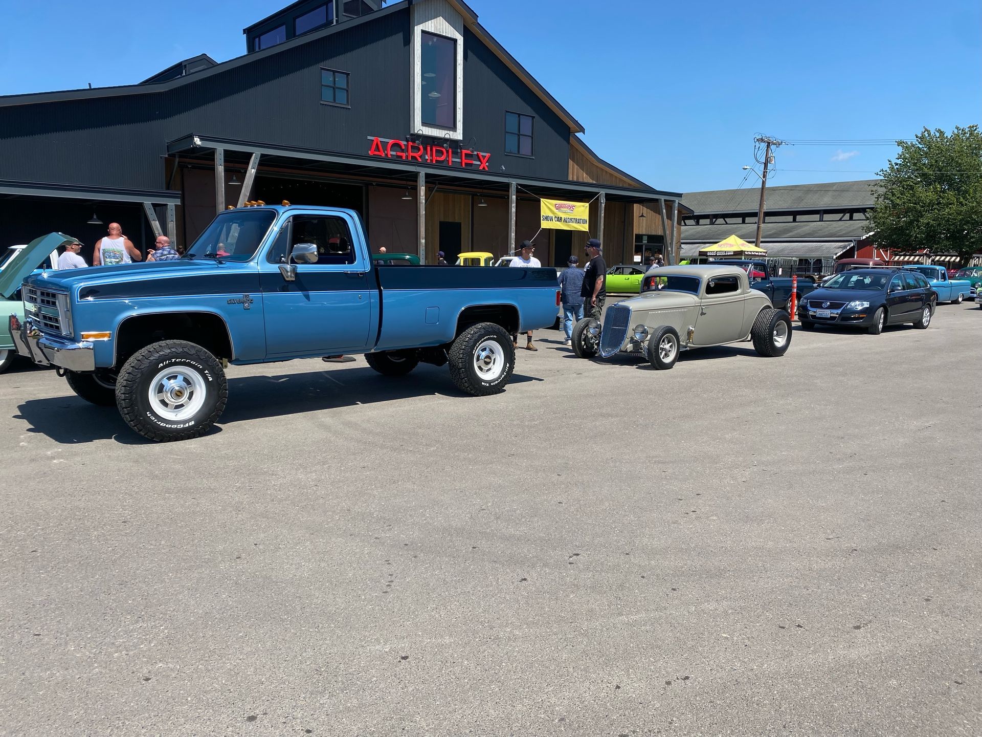 Classic blue pickup and small antique car parked outside a car show building under a clear sky