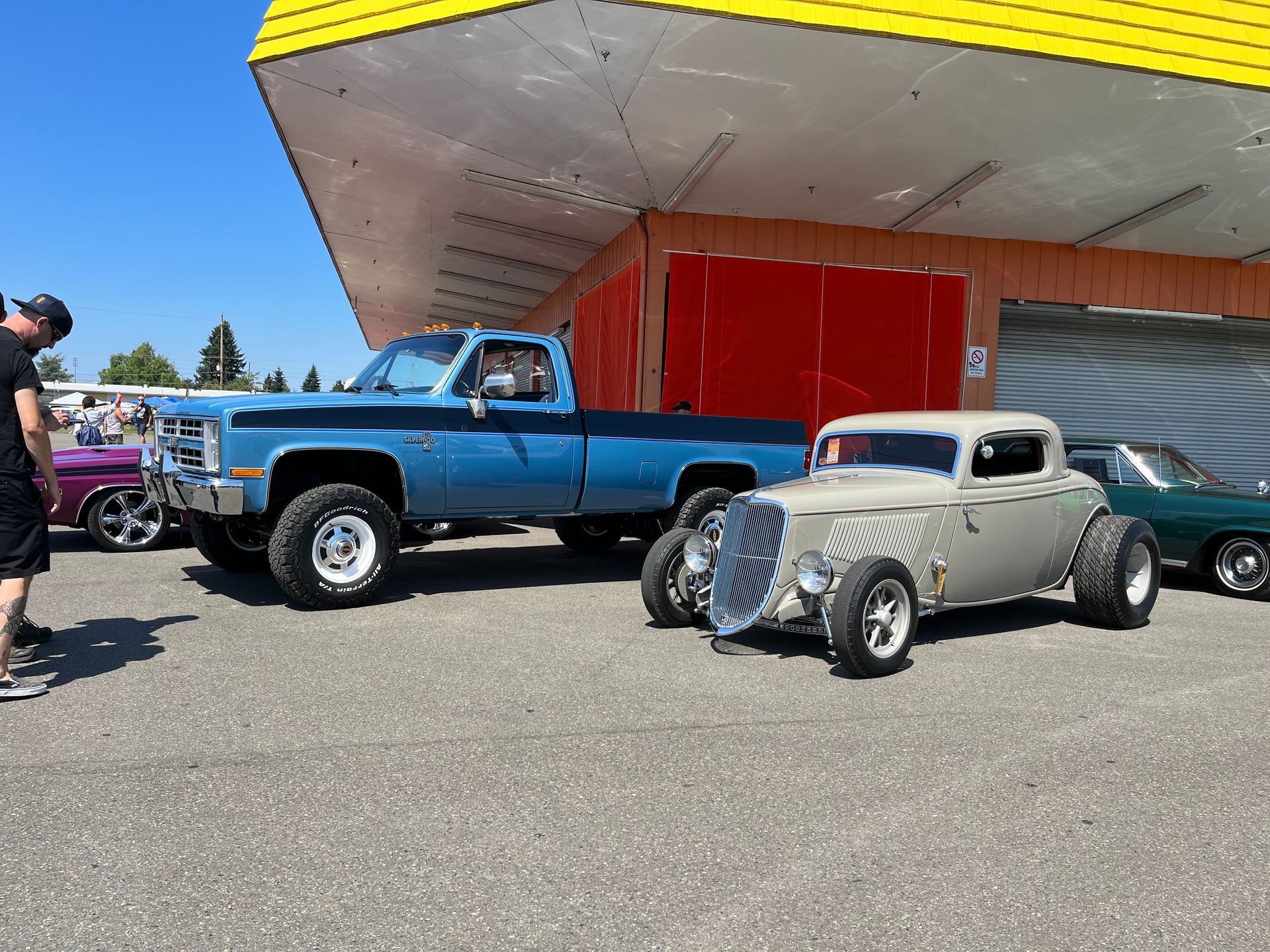 Classic blue pickup and silver hot rod parked at a car show under a red canopy.