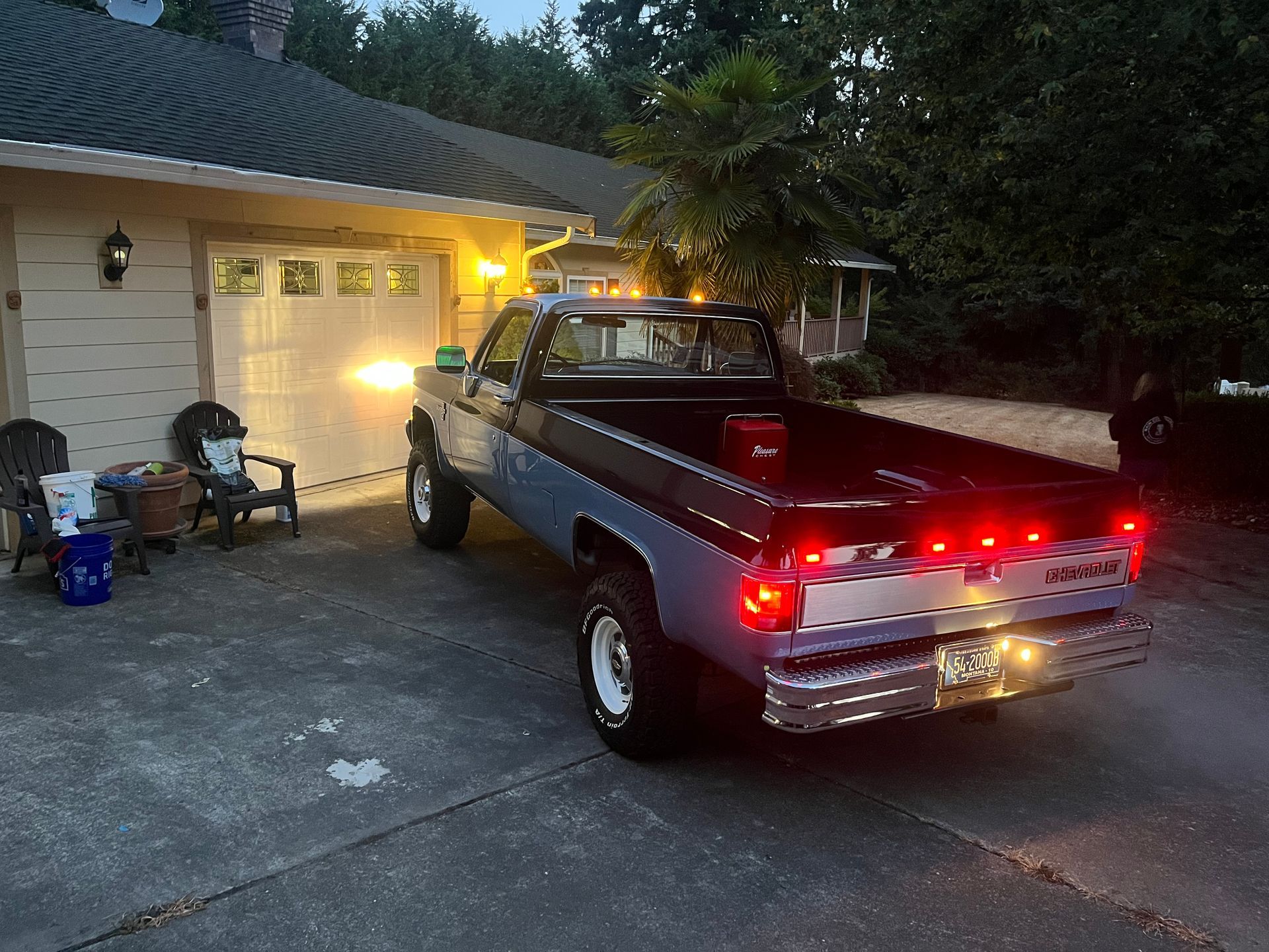 Vintage black pickup truck parked in a driveway at dusk beside a lit garage and house