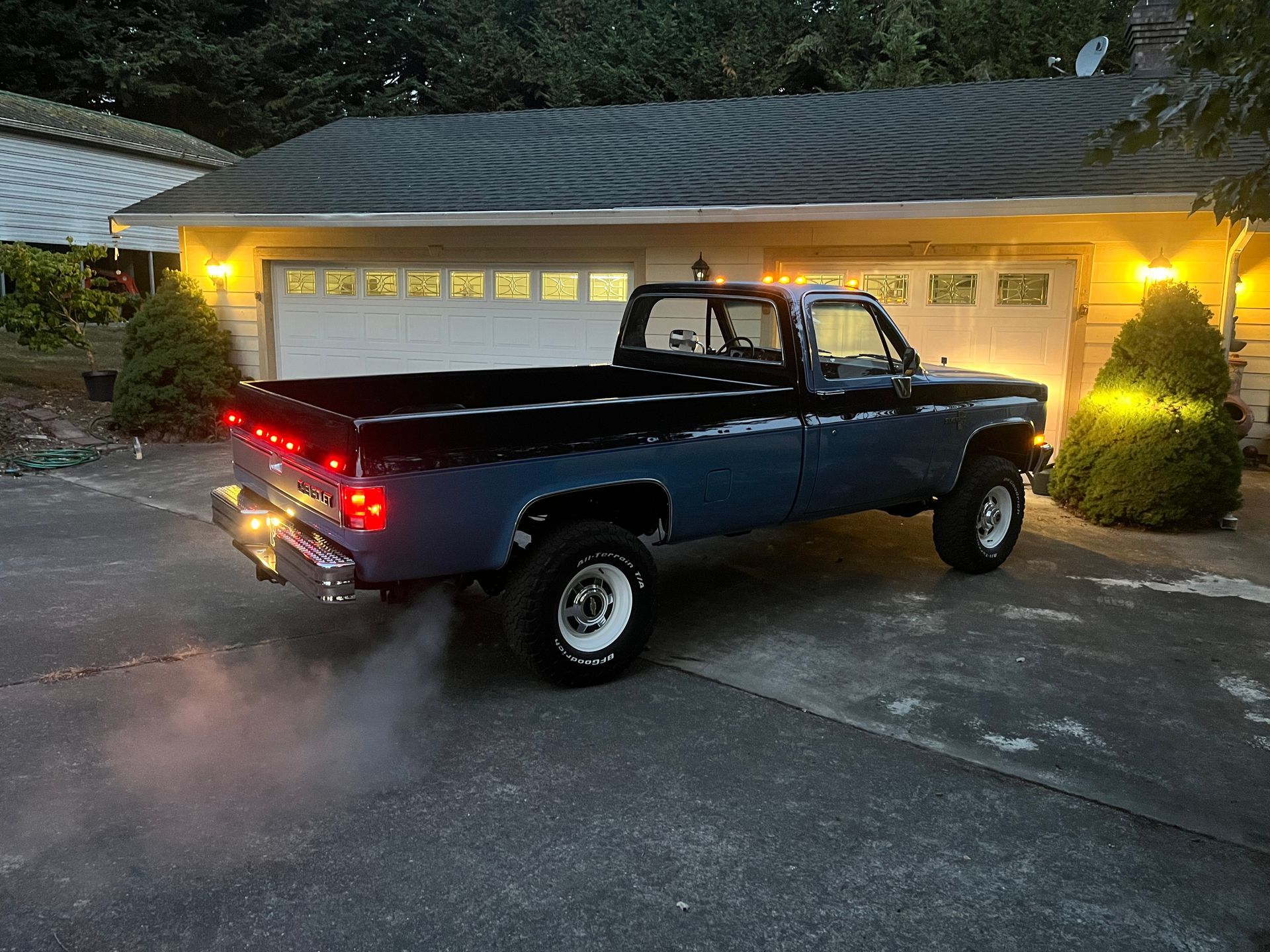 Black pickup truck with smoke from exhaust parked at dusk in front of a lit garage