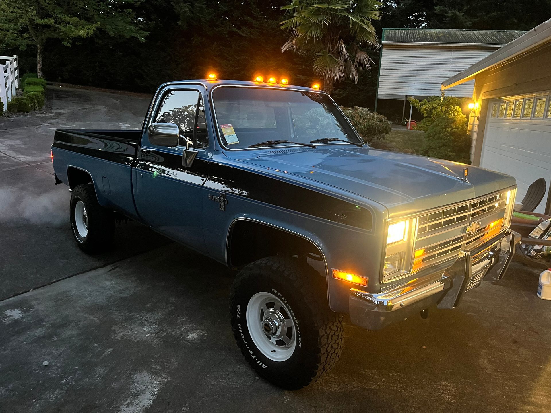 Blue lifted pickup truck with amber roof lights parked in a driveway at dusk