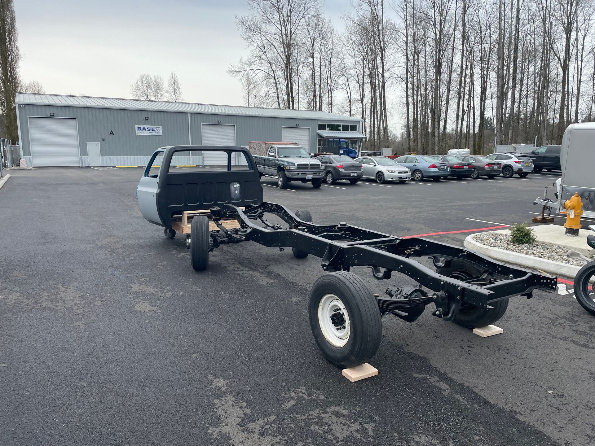 Black truck chassis on blocks in a parking lot beside a gray building and parked cars.