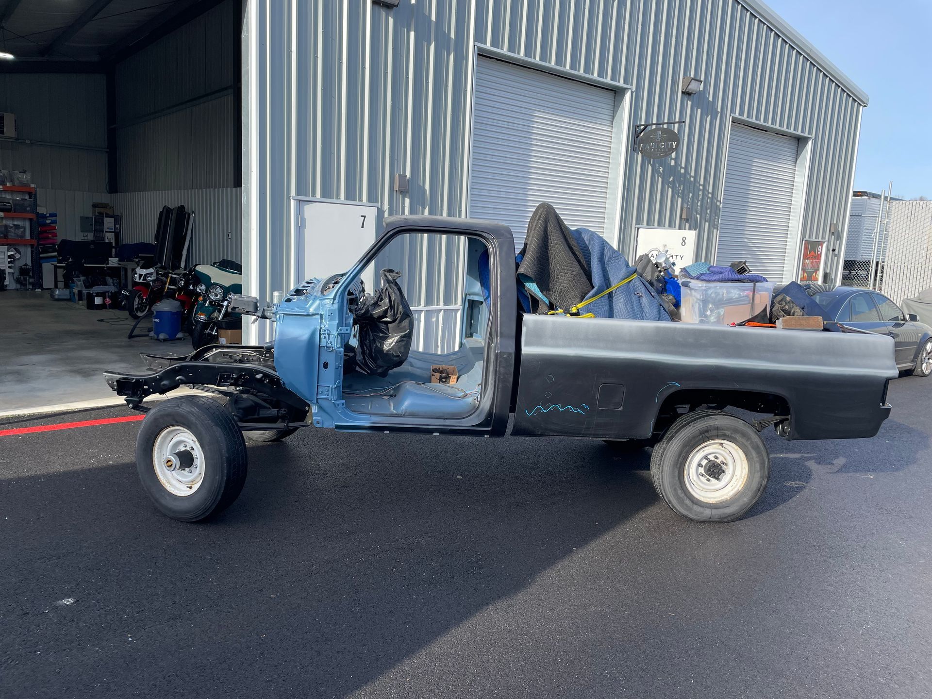 Partially restored blue pickup truck in a warehouse workshop, parked on black asphalt with tools and parts nearby