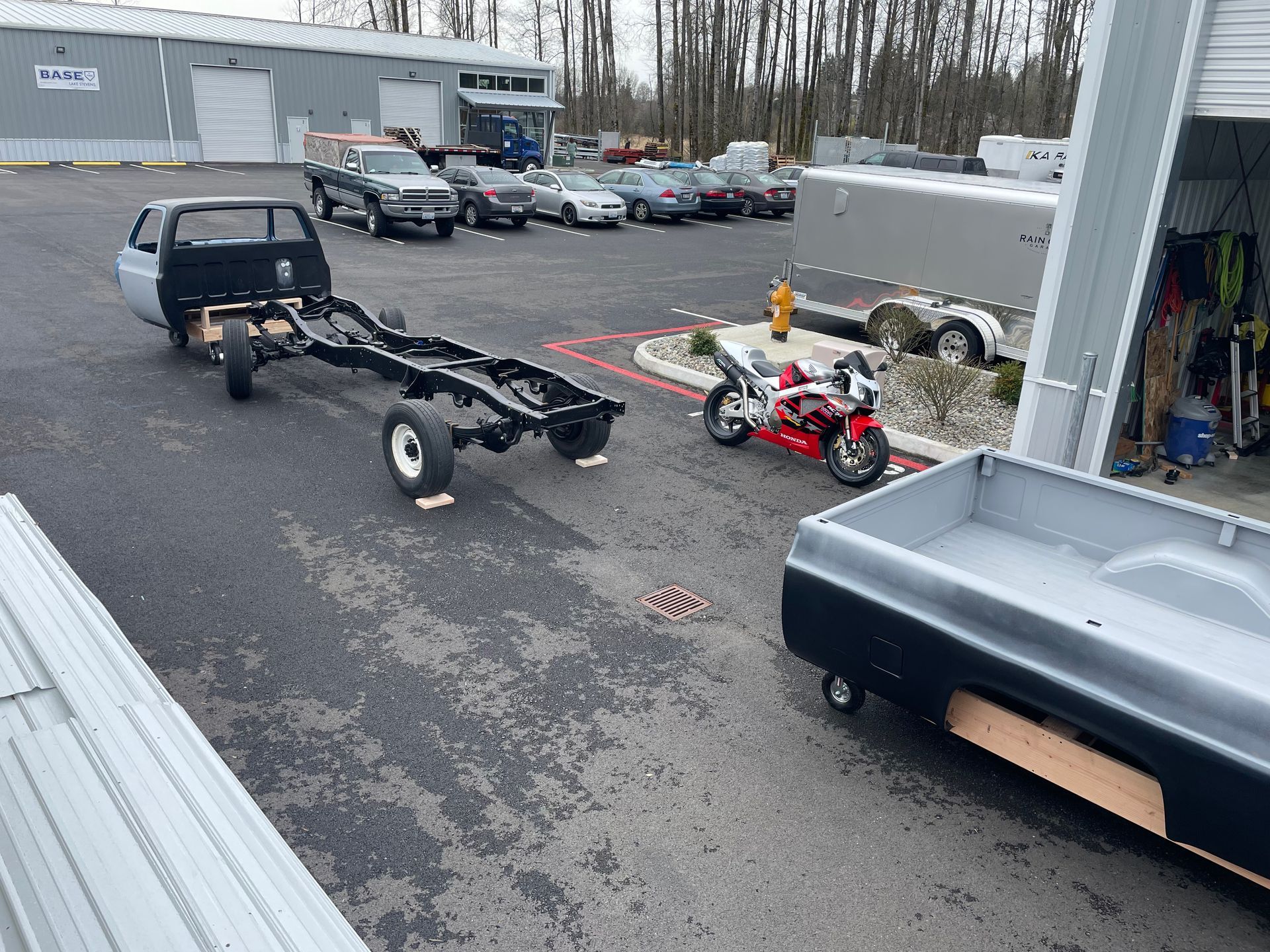 Small trailer and red mini bike parked in an industrial lot beside a building and stacked equipment