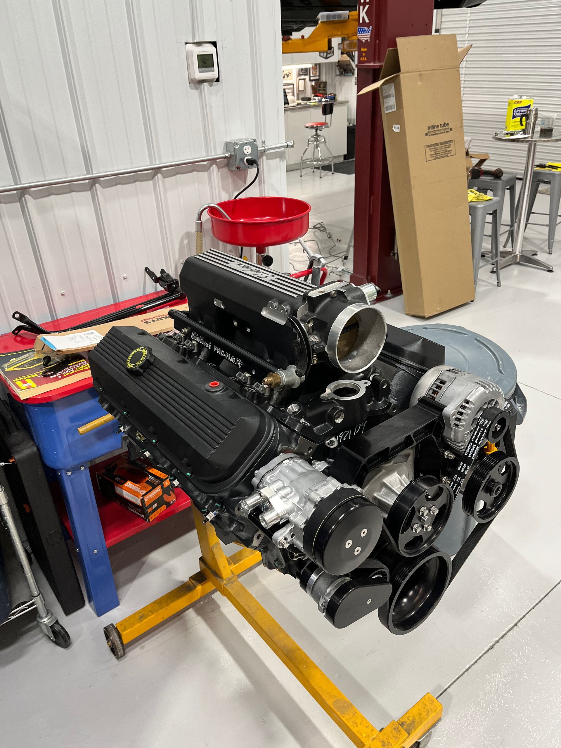 Large car engine on a shop floor beside tools and parts in a garage workshop.