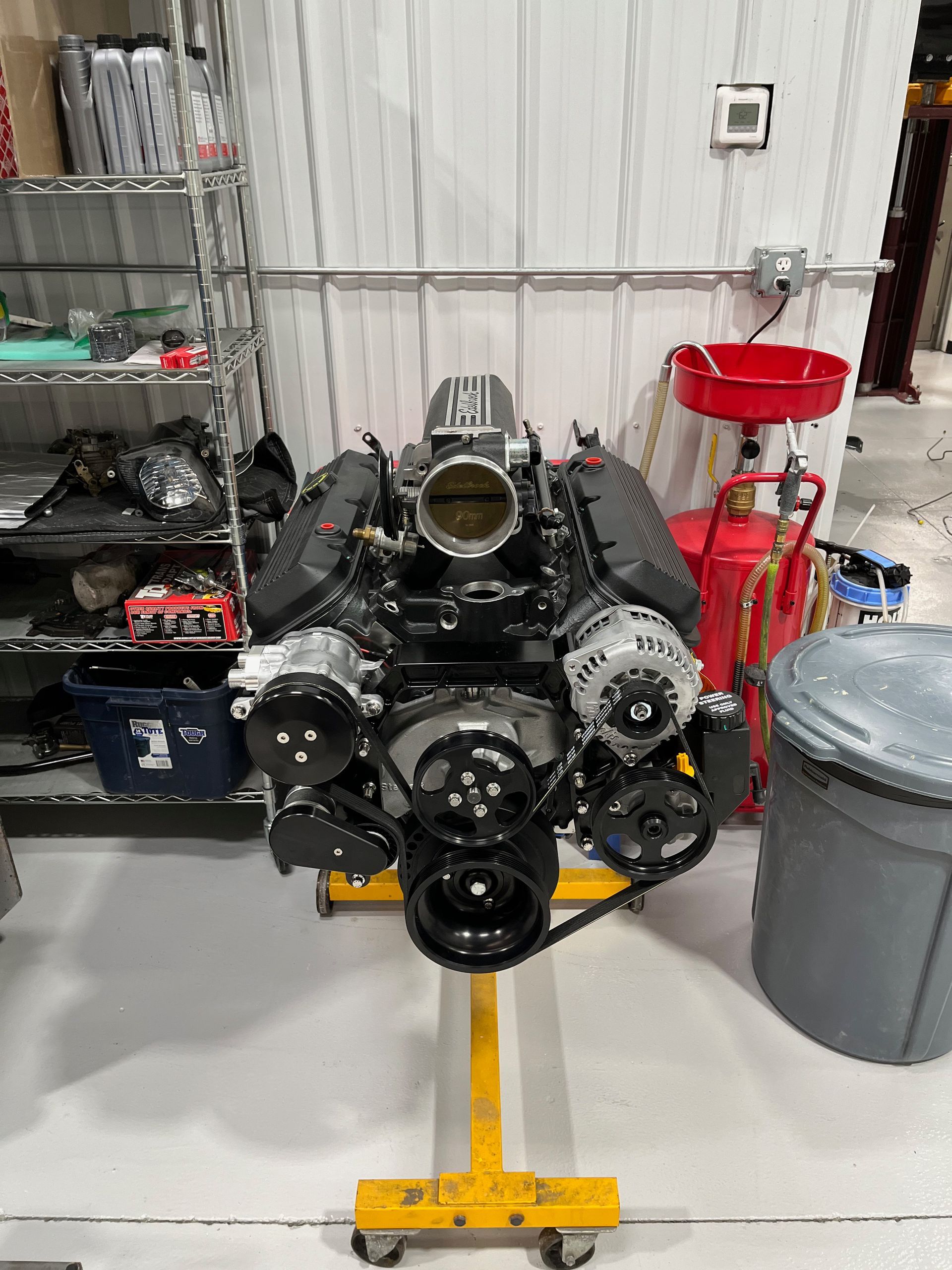 Black engine block on a yellow stand in a workshop, with shelves and a red bin in the background