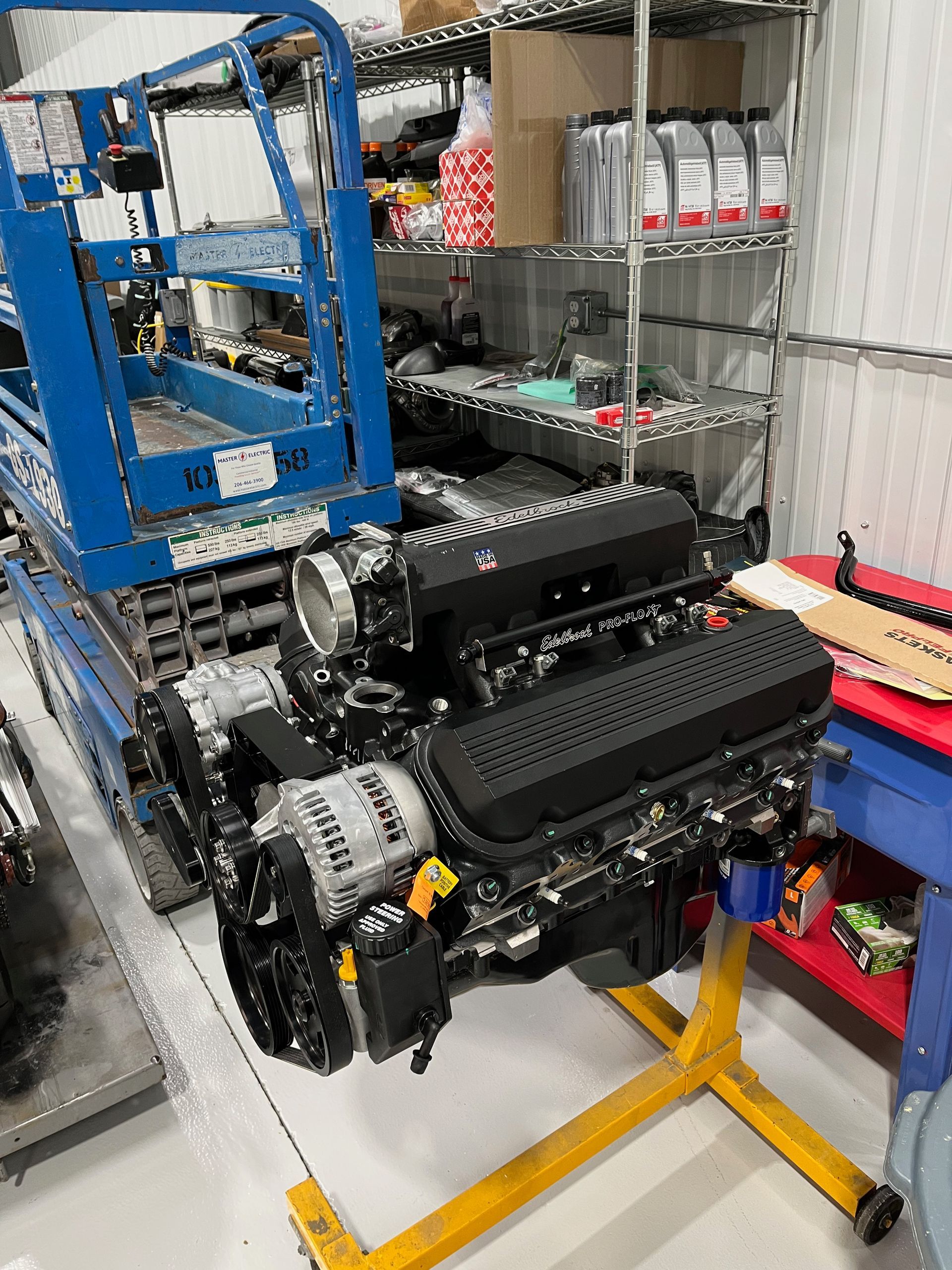 Compact engine assembly on a yellow stand in a workshop, with shelves and a blue cart in the background.