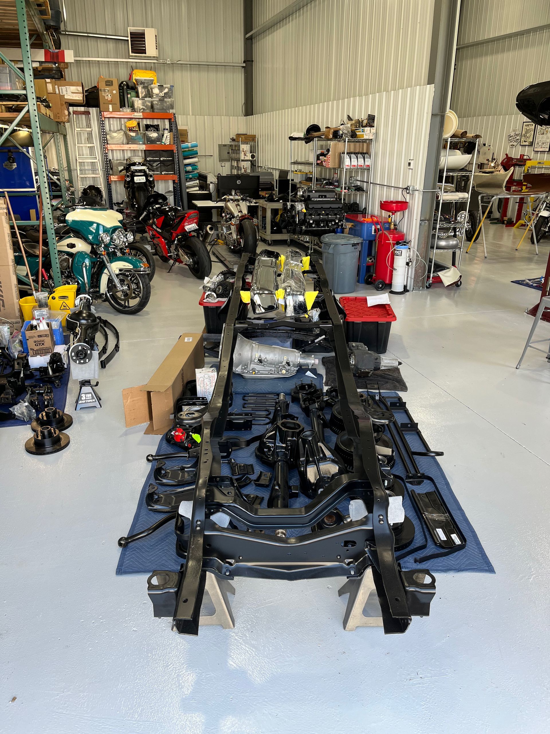 Motorcycle workshop with a stripped chassis centered, surrounded by bikes, tools, and parts on a concrete floor.