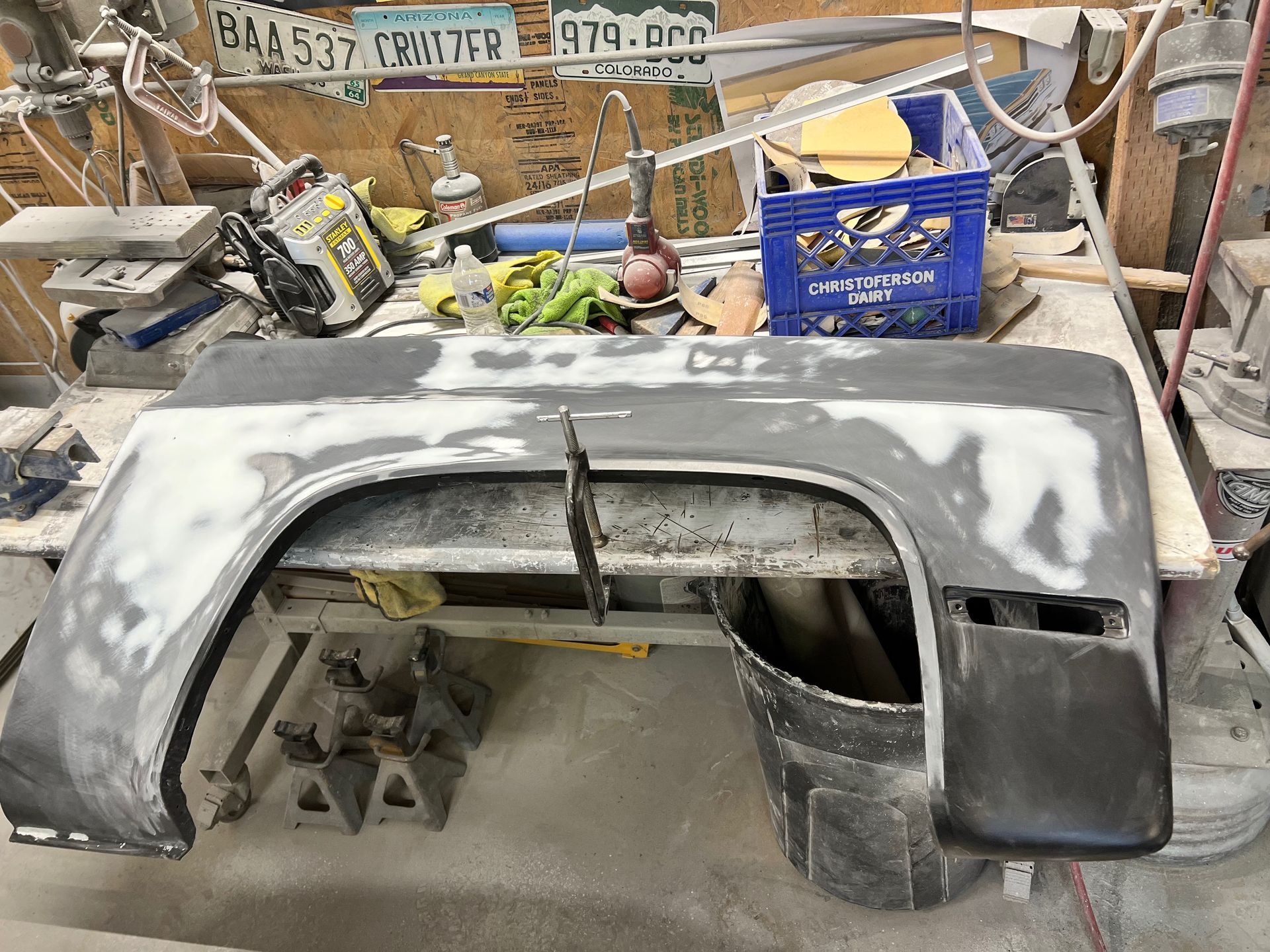 Primered car bumper or body panel resting on a cluttered workbench in a garage.